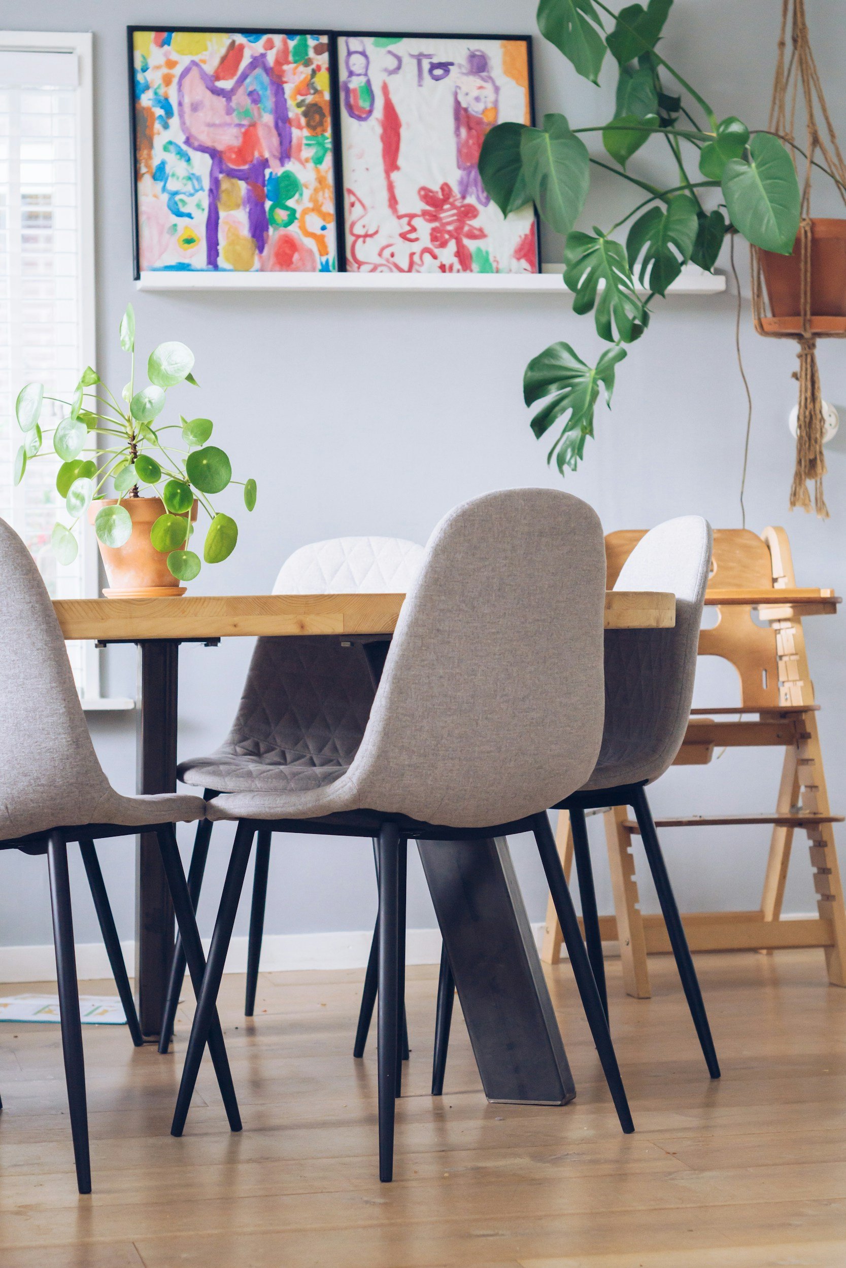 Dining room with a wooden table, beige upholstered chairs, and indoor plants. Two colorful abstract paintings hang on the wall above the shelf, and a high chair can be seen in the background.
