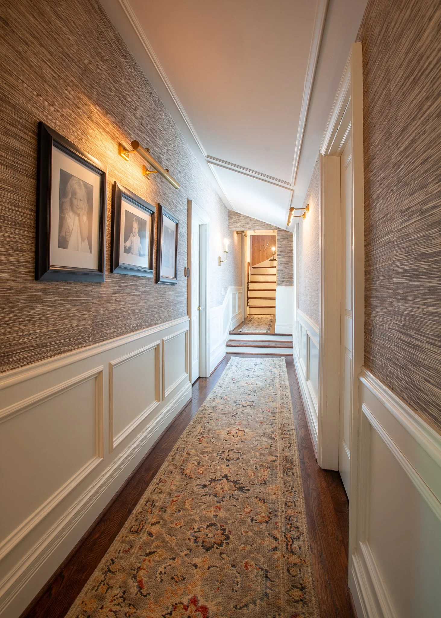 A hallway with wood flooring, beige wainscoting, textured wallpaper, and framed photographs on the wall. It has multiple wall-mounted lights and a patterned runner rug on the floor. The hallway leads to a staircase.
