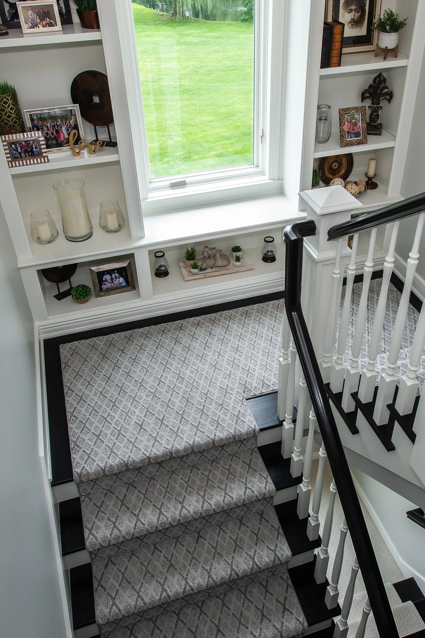 View of a staircase with carpeted steps leading down to a landing, featuring a white banister with black handrails, and a built-in shelf with framed photographs and decorative items, next to a window showing a view of a grassy yard.