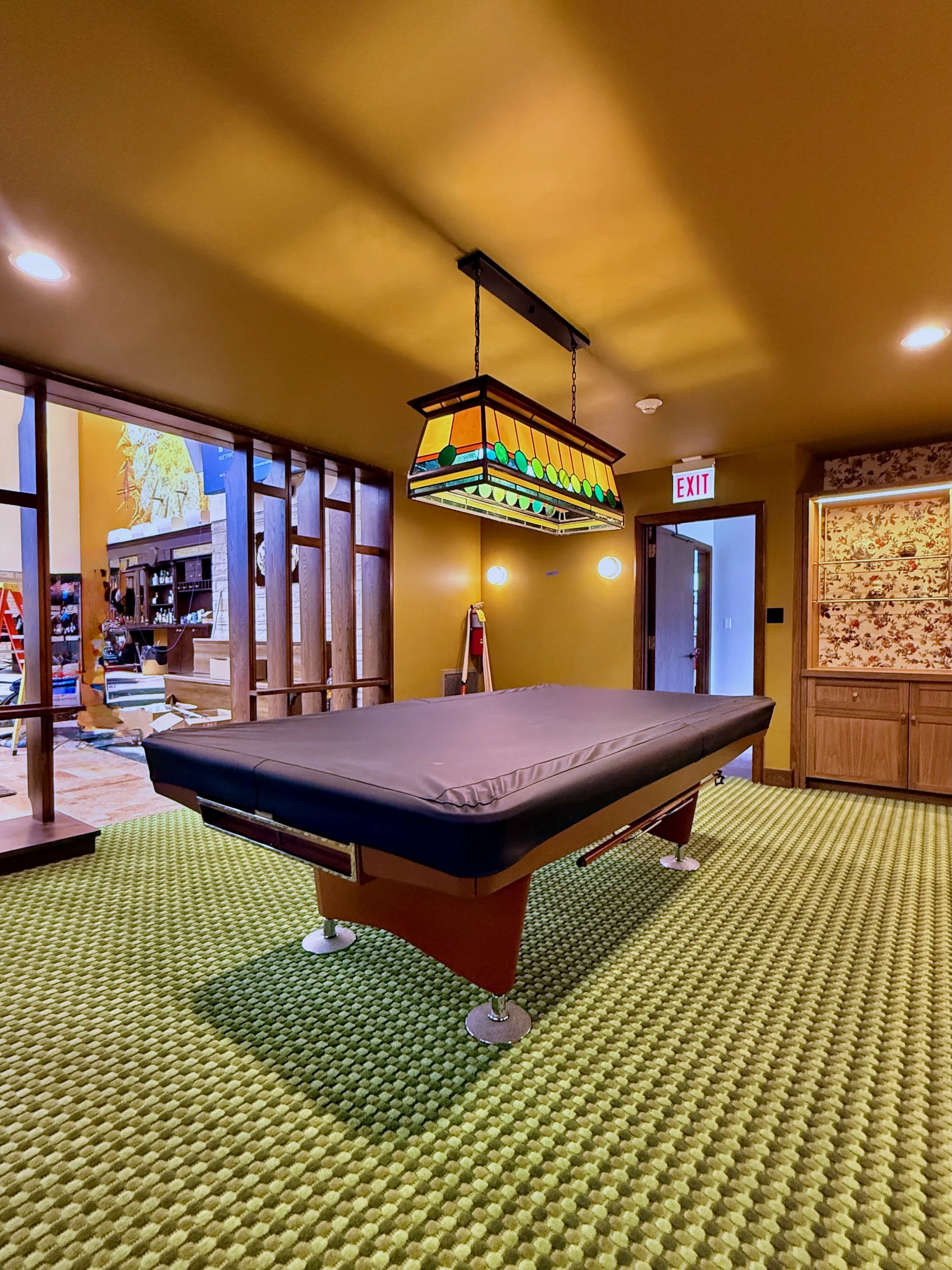An indoor recreational room featuring a black shuffleboard table on a green patterned carpet, a colorful stained glass ceiling light, and warm yellow walls. There are wood and glass partitions, and a door with a red EXIT sign in the background.