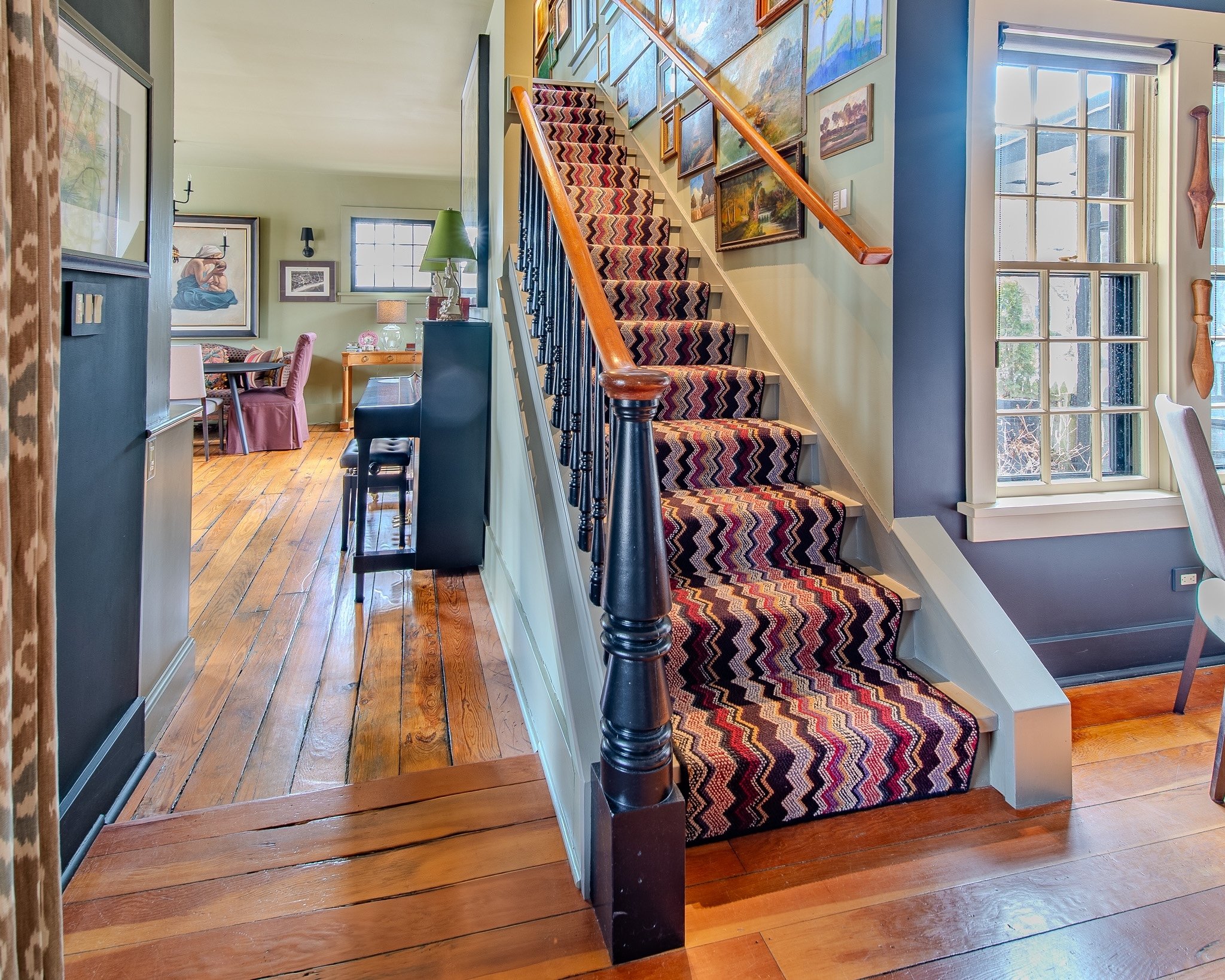 Interior view of a house showing a staircase with a colorful zigzag patterned carpet, a wooden banister, and framed paintings on the wall. There are large windows allowing natural light into the space, and a hardwood floor extending throughout the ar