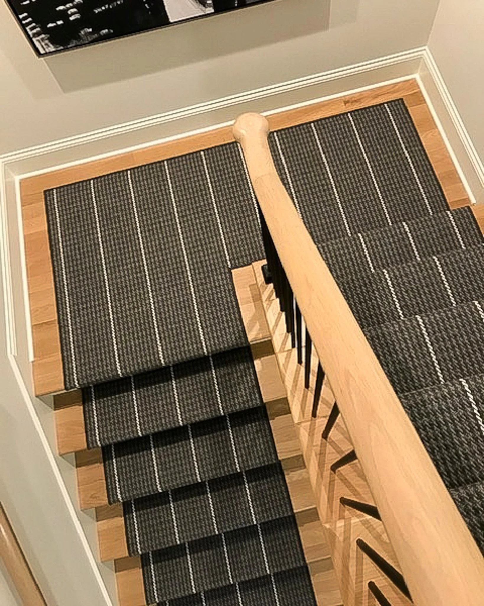 From the top of the staircase, looking down shows wooden stairs with black and white striped carpet runners, a blackboard or poster on the gray wall at the top of the stairs, and a wooden handrail along the staircase.