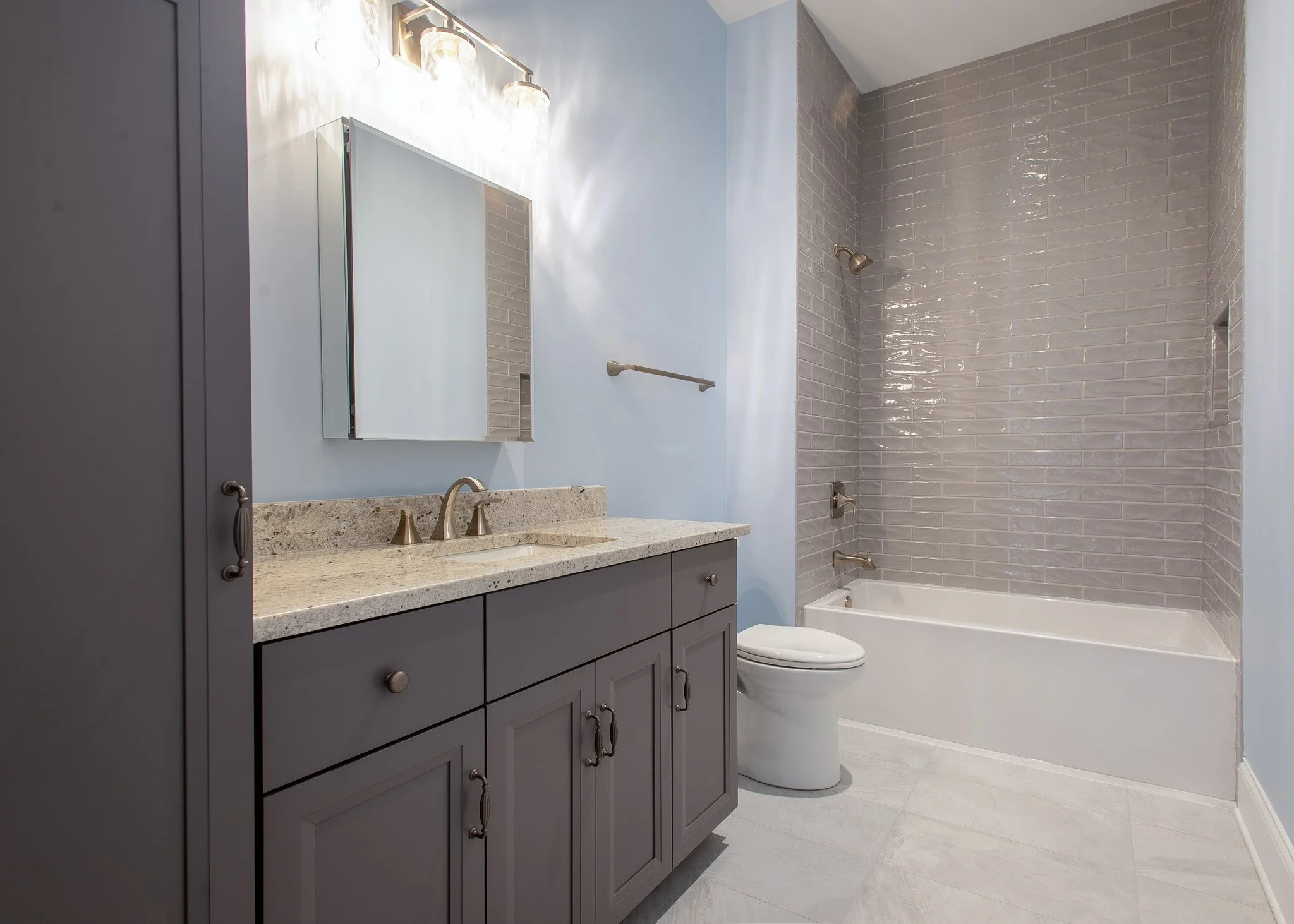 A modern bathroom with a blue wall, gray vanity with multiple drawers, a beige stone countertop, and a mirror. There is a white toilet next to a bathtub with beige subway tile walls, a shower head, and a grab bar. The floor is light-colored tile.