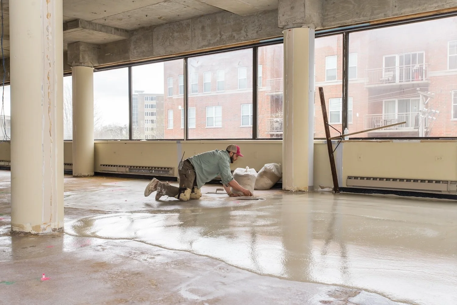 A construction worker kneeling on a wet floor inside a building, smoothing or preparing concrete. Large windows overlooking neighboring buildings are in the background.