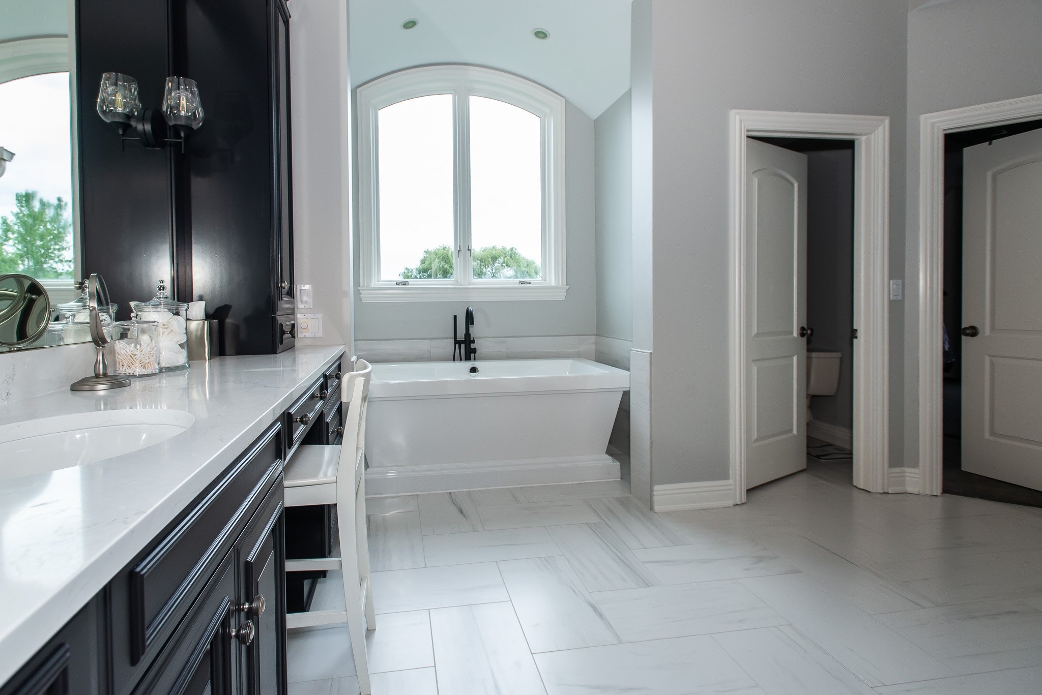 Bright bathroom with white marble floor, large window, and a freestanding bathtub. Black cabinetry with glassware on the counter and two doorways, one leading to a toilet room.