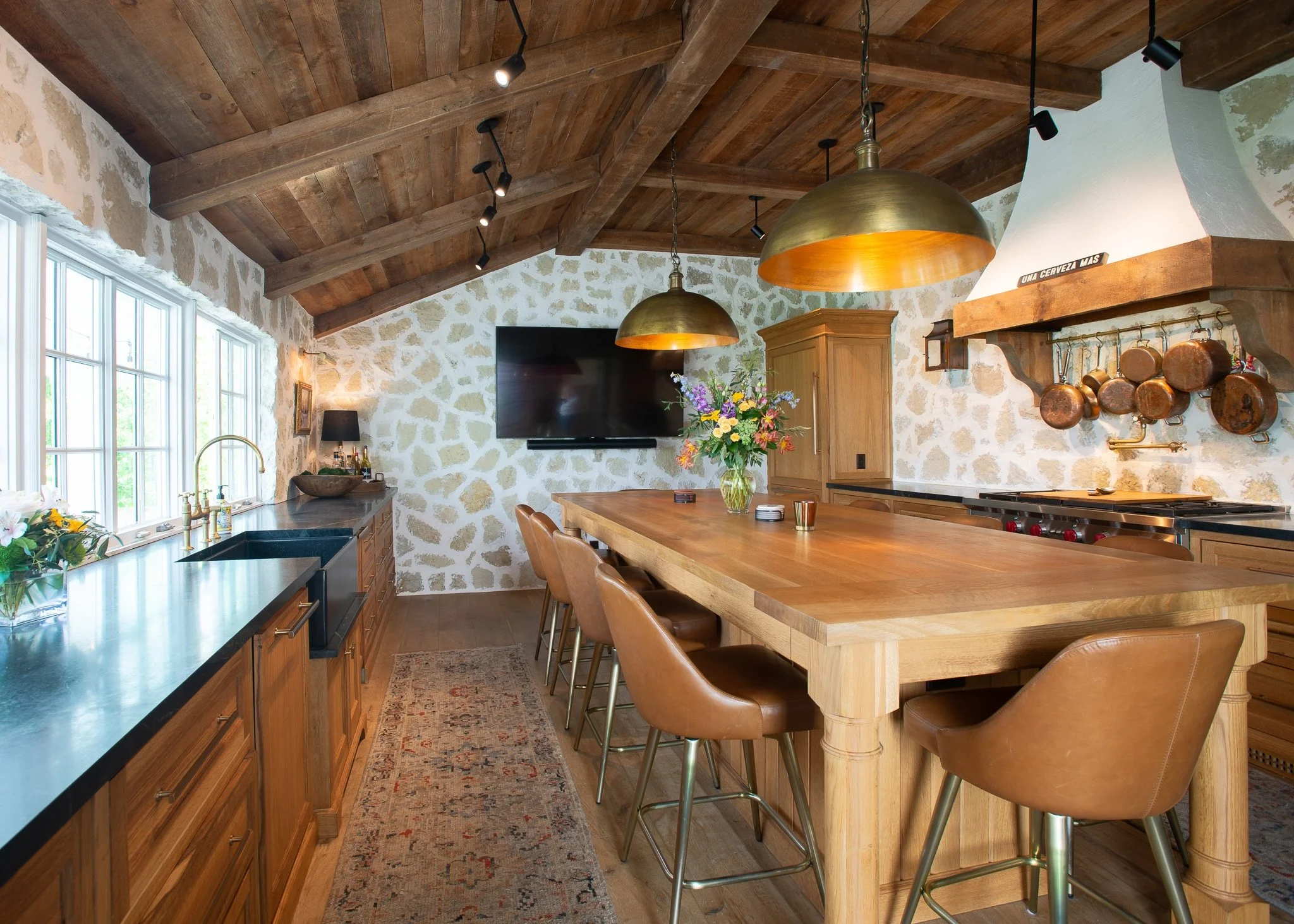 A rustic kitchen with a wooden ceiling and stone walls, featuring a large wooden dining table, leather chairs, brass pendant lights, a stone fireplace mantel with hanging copper pots, a large TV on the wall, and kitchen cabinets with a vase of flower