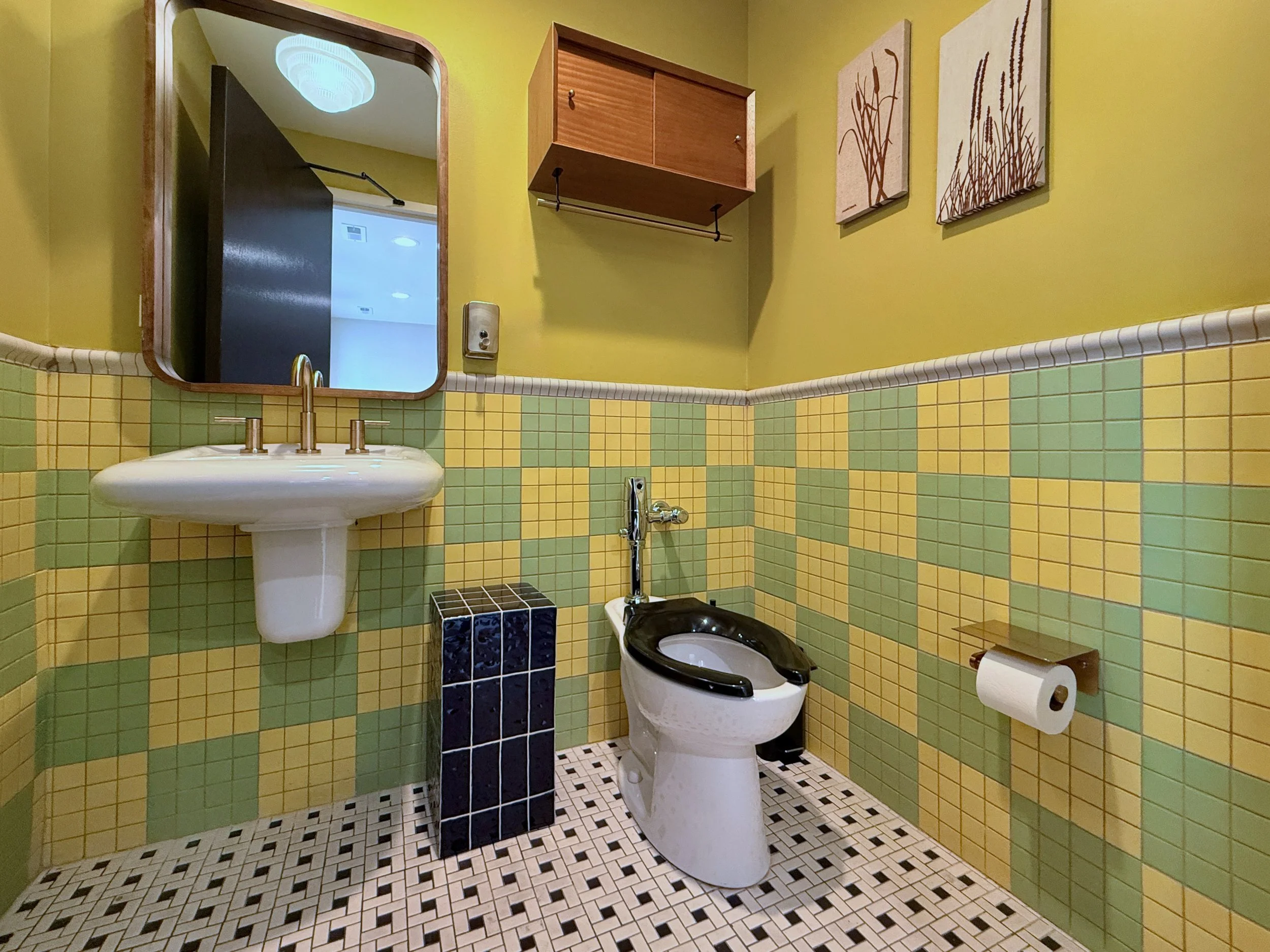 Small bathroom with yellow and green tiled walls, a white sink with gold faucet, a black and white toilet, a mirror above the sink, a small black cubical decor piece, wall-mounted cabinet, art pieces with plant designs, and a roll of toilet paper.