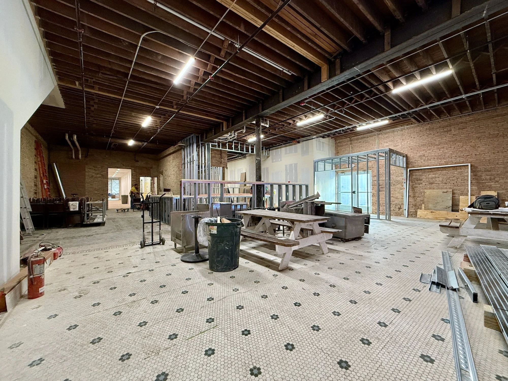 Interior of a building under construction or renovation with exposed brick walls, a partially finished ceiling with visible wooden beams and lighting fixtures, construction materials, tools, and work tables scattered across the space.