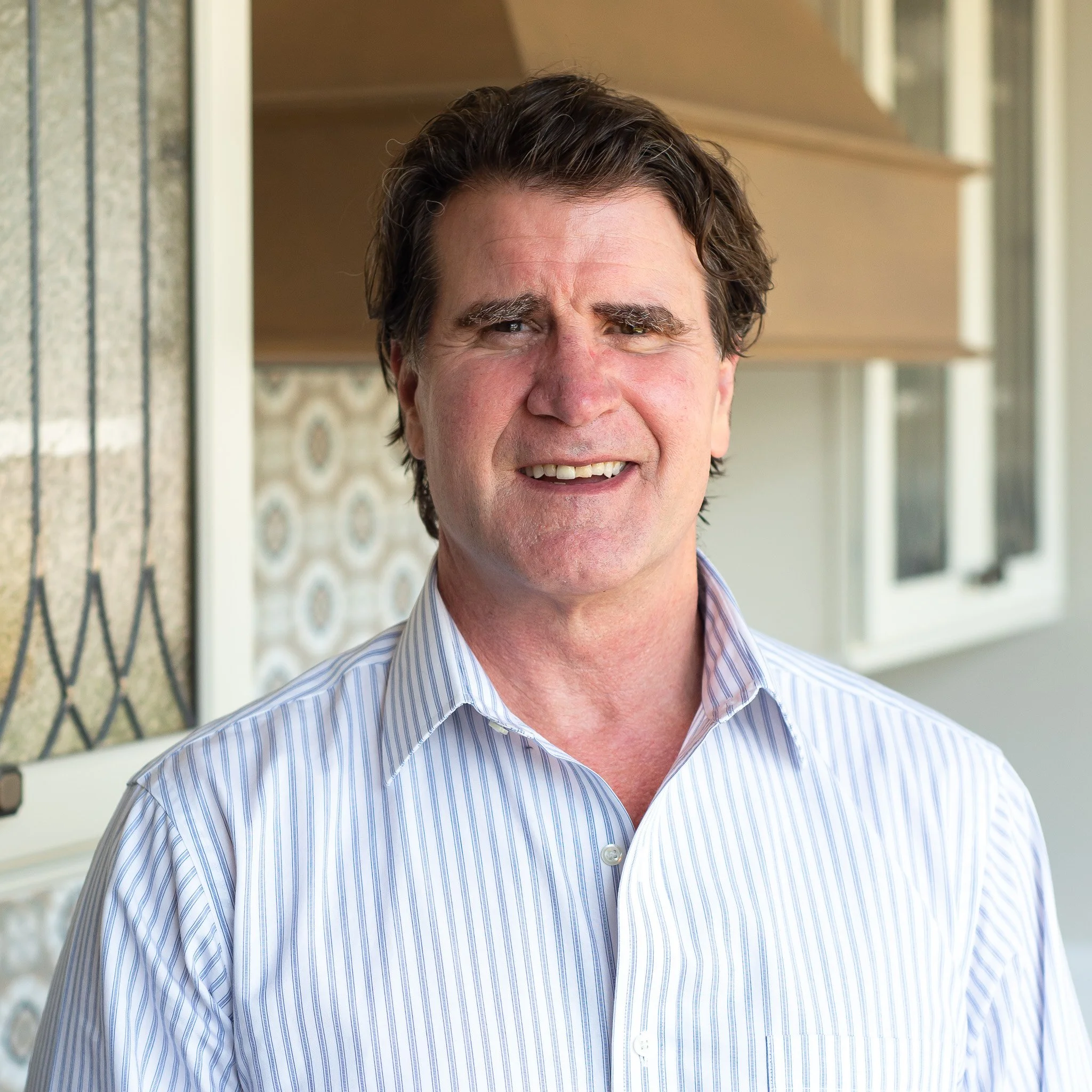 A middle-aged man with dark, wavy hair and a big smile, wearing a light blue and white striped shirt, standing outside near a house with a screened door and patterned decorative tiles.