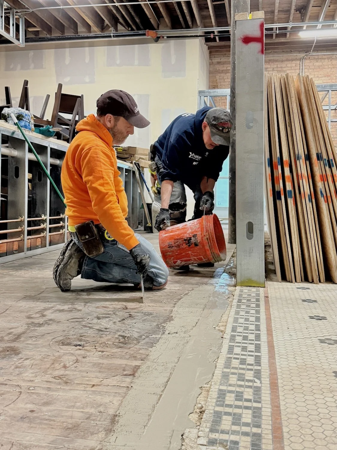 Two construction workers kneel on the floor, pouring and spreading a cement mix along the edge of a tiled area in an unfinished interior space with construction materials, tools, and stacked wood around.