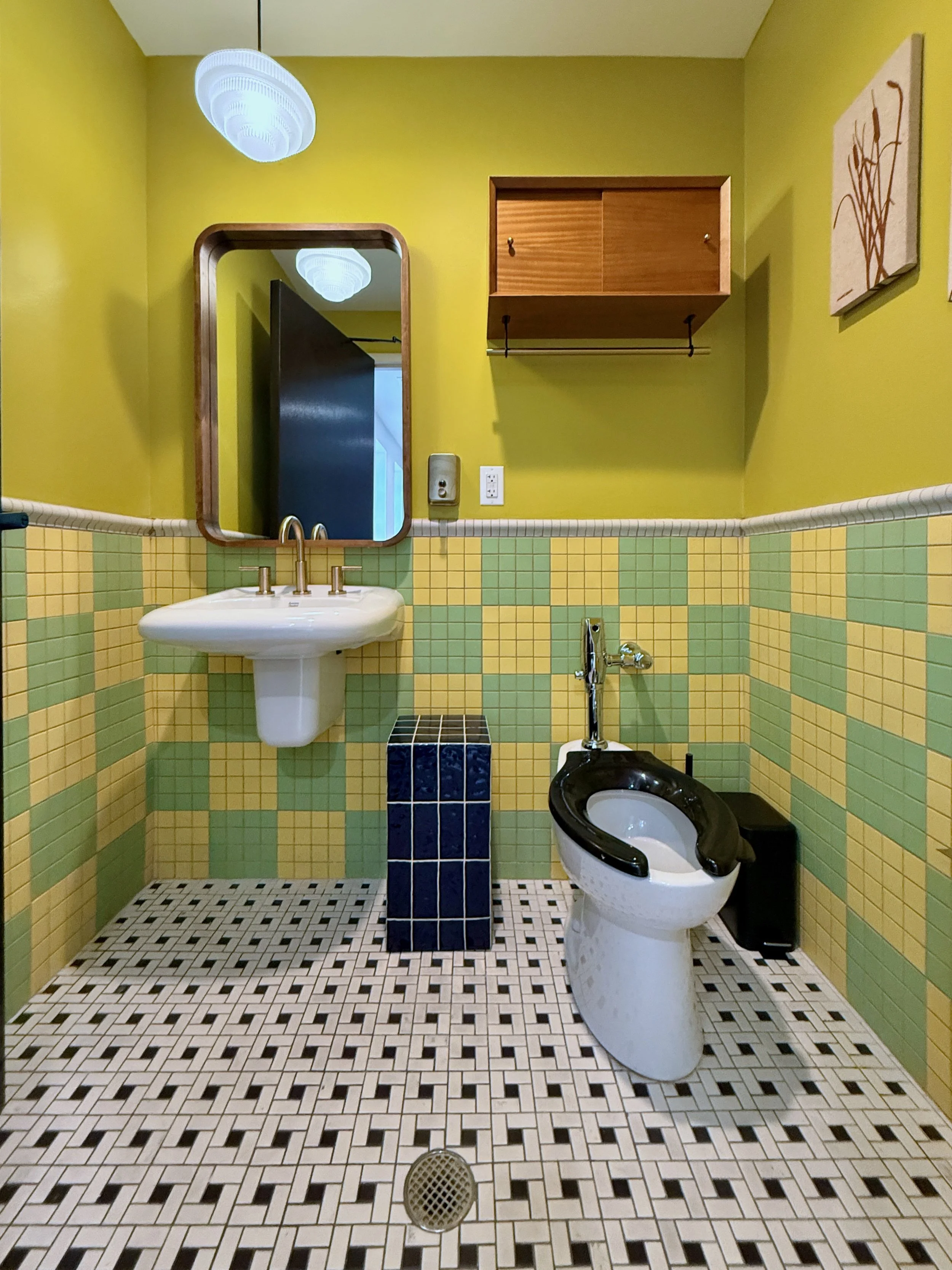 A vintage-style bathroom with yellow walls, green and yellow tiled wainscoting, checkered black and white floor tiles, a small white sink with a gold faucet, a black and white toilet, a wall-mounted wooden cabinet, a mirror, and a ceiling light fixtu