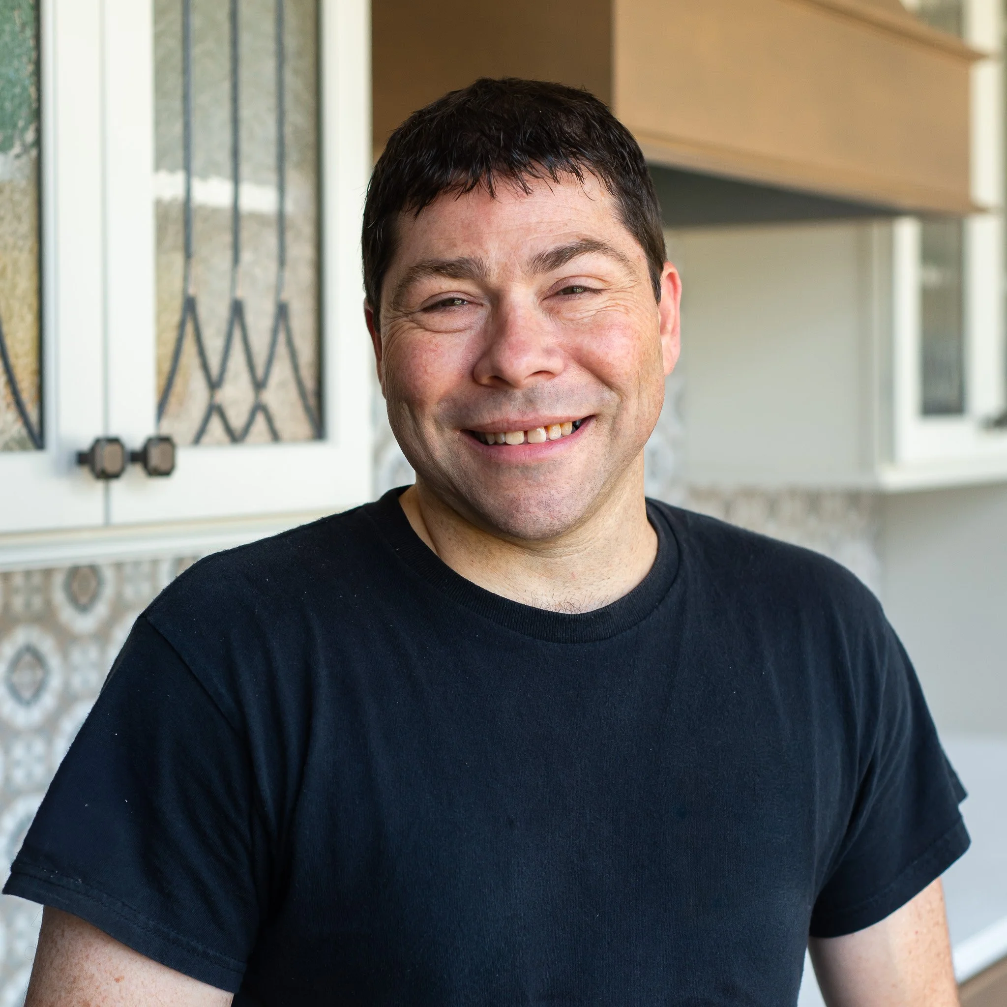 Close-up of a smiling person with short dark hair wearing a black t-shirt, standing in front of a window with patterned glass and a beige wall.