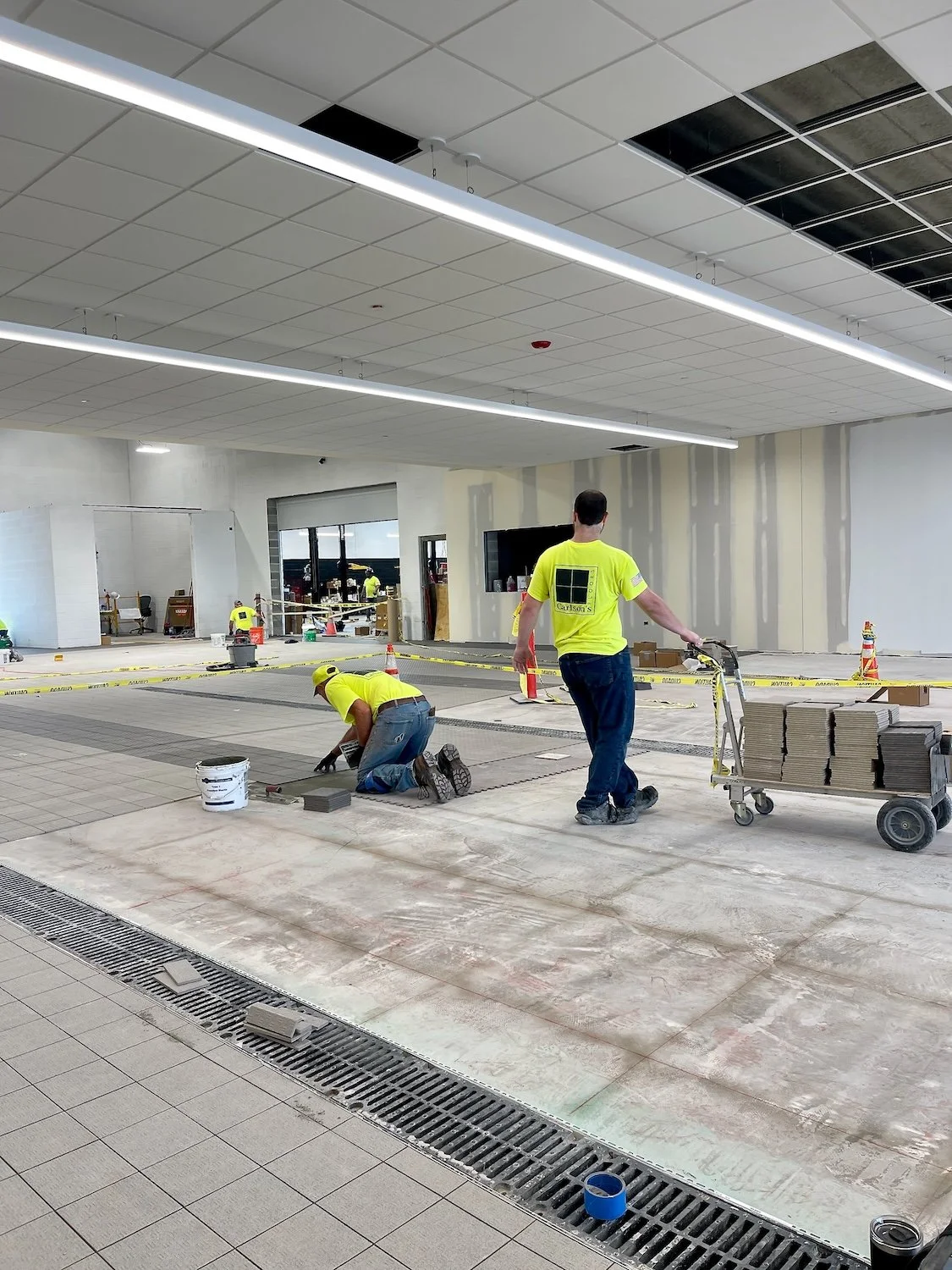 Construction workers installing floor tiles in an indoor space, with some workers kneeling and others pushing a cart filled with tiles.