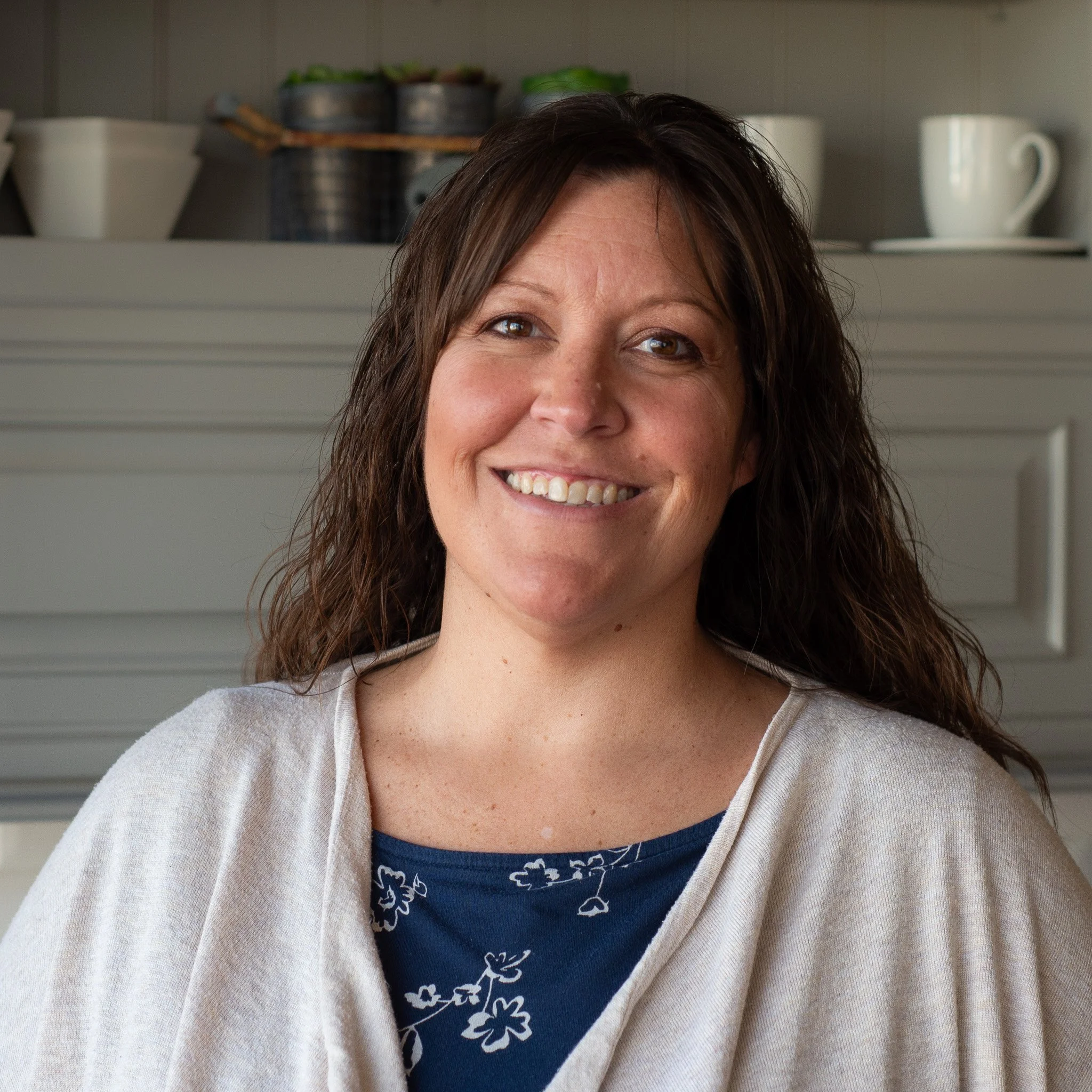 Close-up of a smiling woman with brown hair, wearing a navy blue shirt with white floral patterns and a light-colored cardigan, standing in front of a shelf with cups and containers.