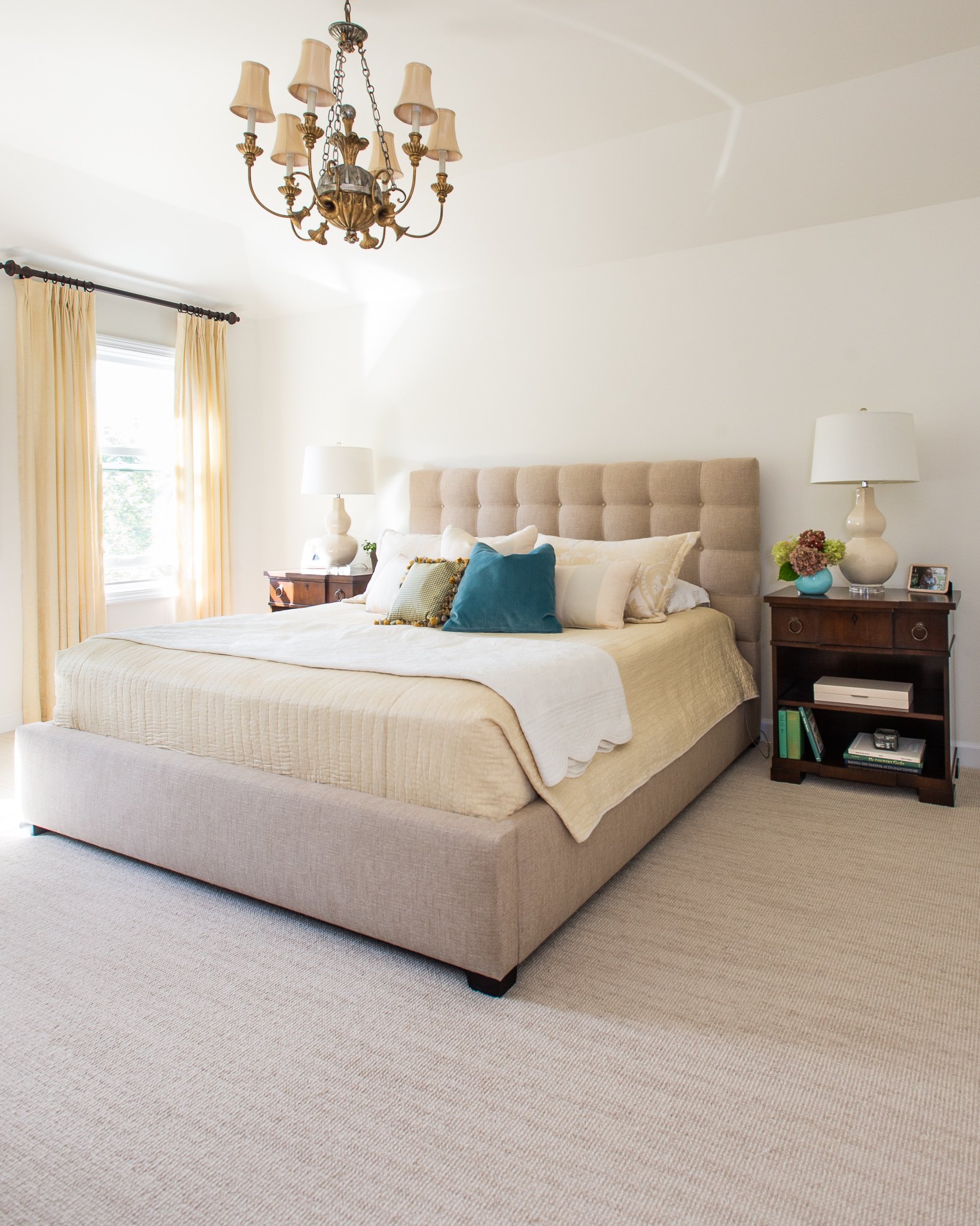 A bedroom with cream-colored bedding, a beige upholstered headboard, and multiple pillows. A wooden nightstand holds a white lamp, a small framed photo, and a vase with flowers. Yellow curtains are on a window, and a brass chandelier hangs from the c
