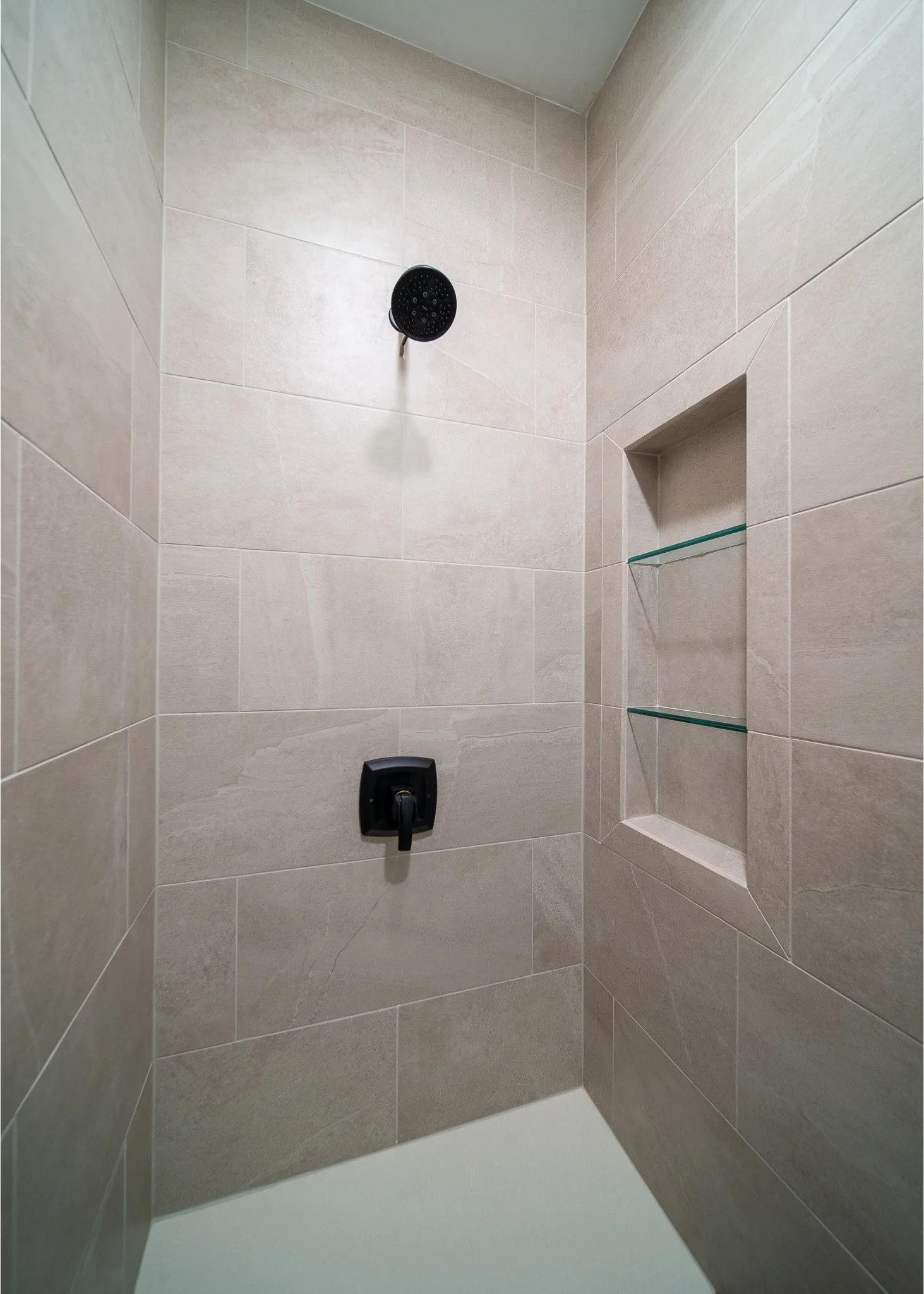 A shower area with beige tiled walls, a black shower head, a black control valve, and three glass shelves built into the wall.
