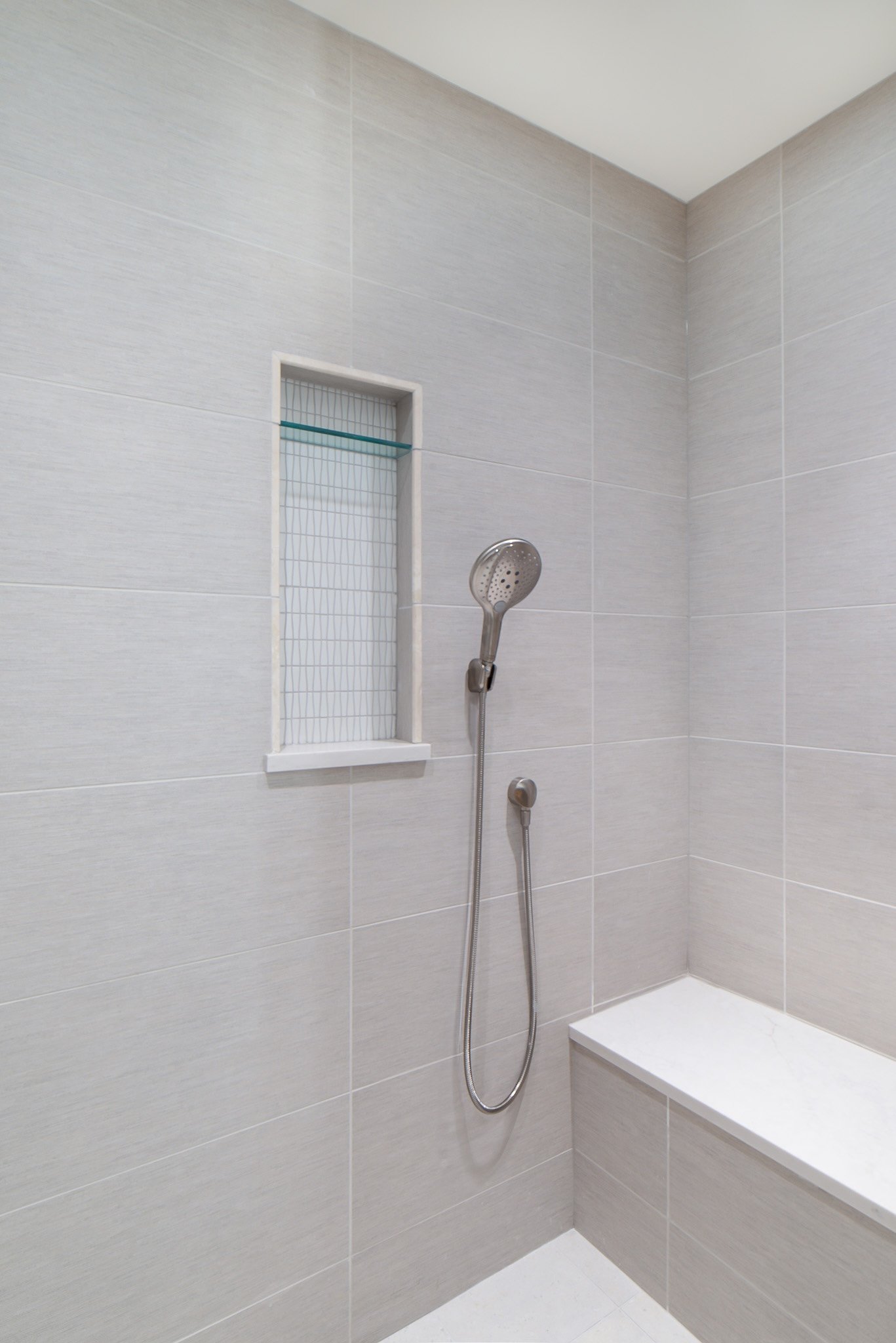 Shower area with light gray tiled walls, a handheld showerhead, a small built-in niche with a glass shelf, and a marble bench in the corner.