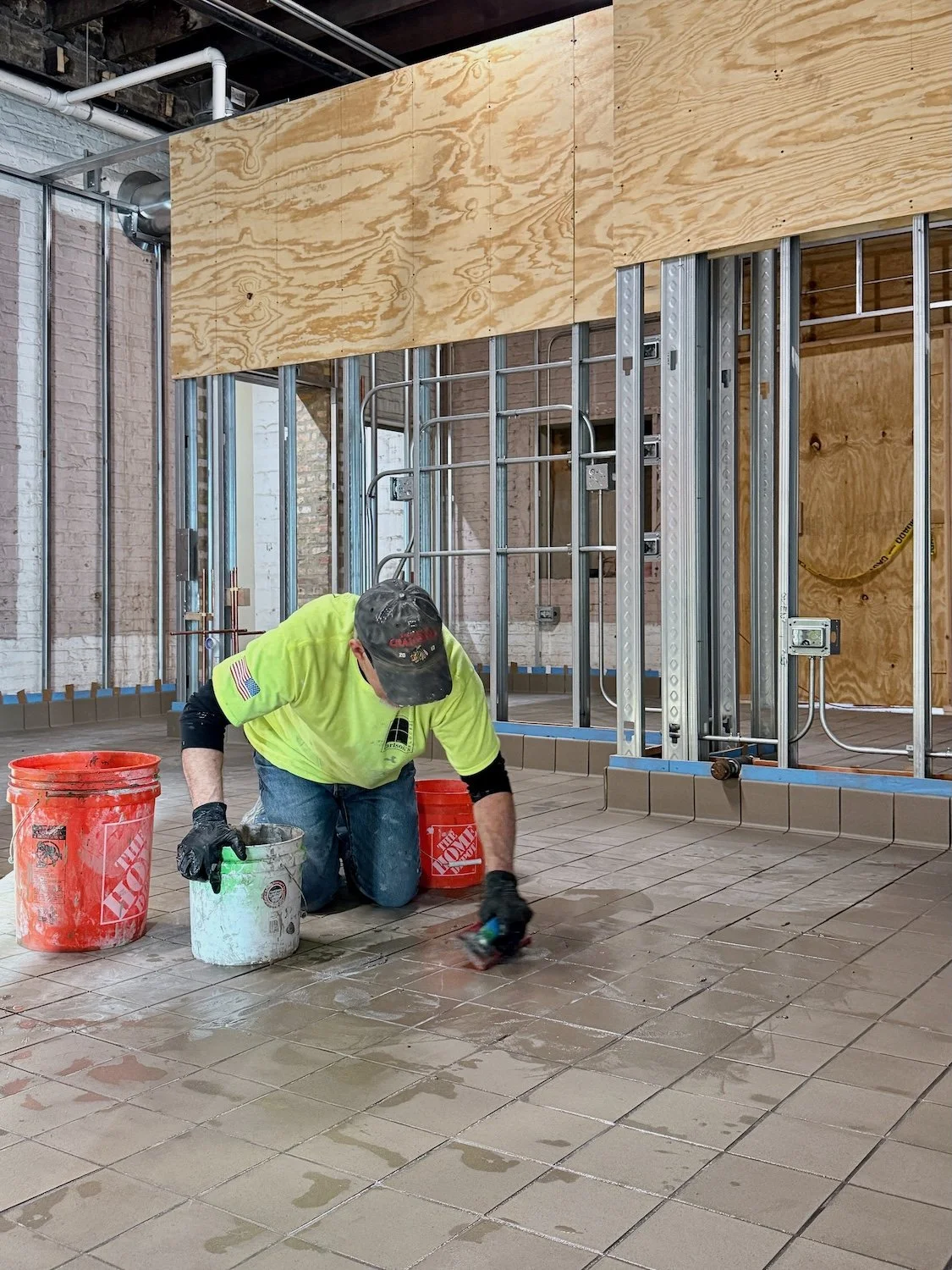 A construction worker kneeling on the floor working on tile installation at a building site with exposed wall framing and plywood panels.