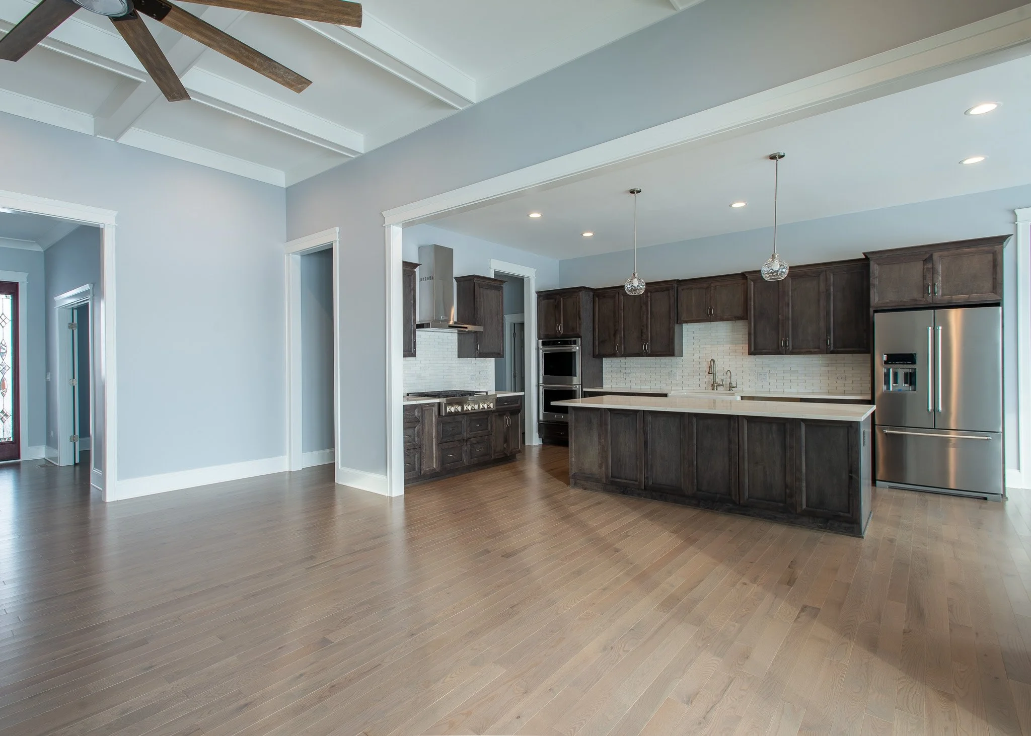 A modern kitchen with dark wooden cabinets, white backsplash, stainless steel appliances, pendant lights, and a light wood floor.