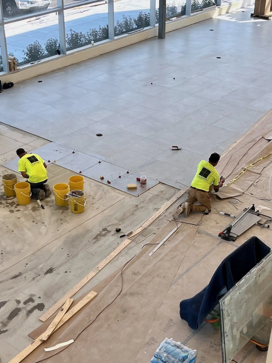 Two construction workers install large gray tiles on the floor of a building's interior, with construction tools and materials around them.