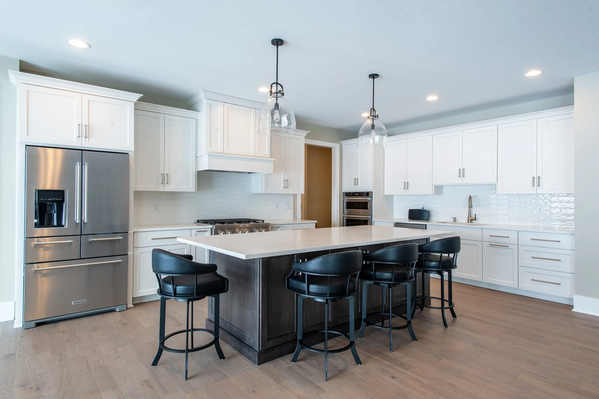 Modern white kitchen with an island, black chairs, stainless steel refrigerator, oven, and pendant lighting