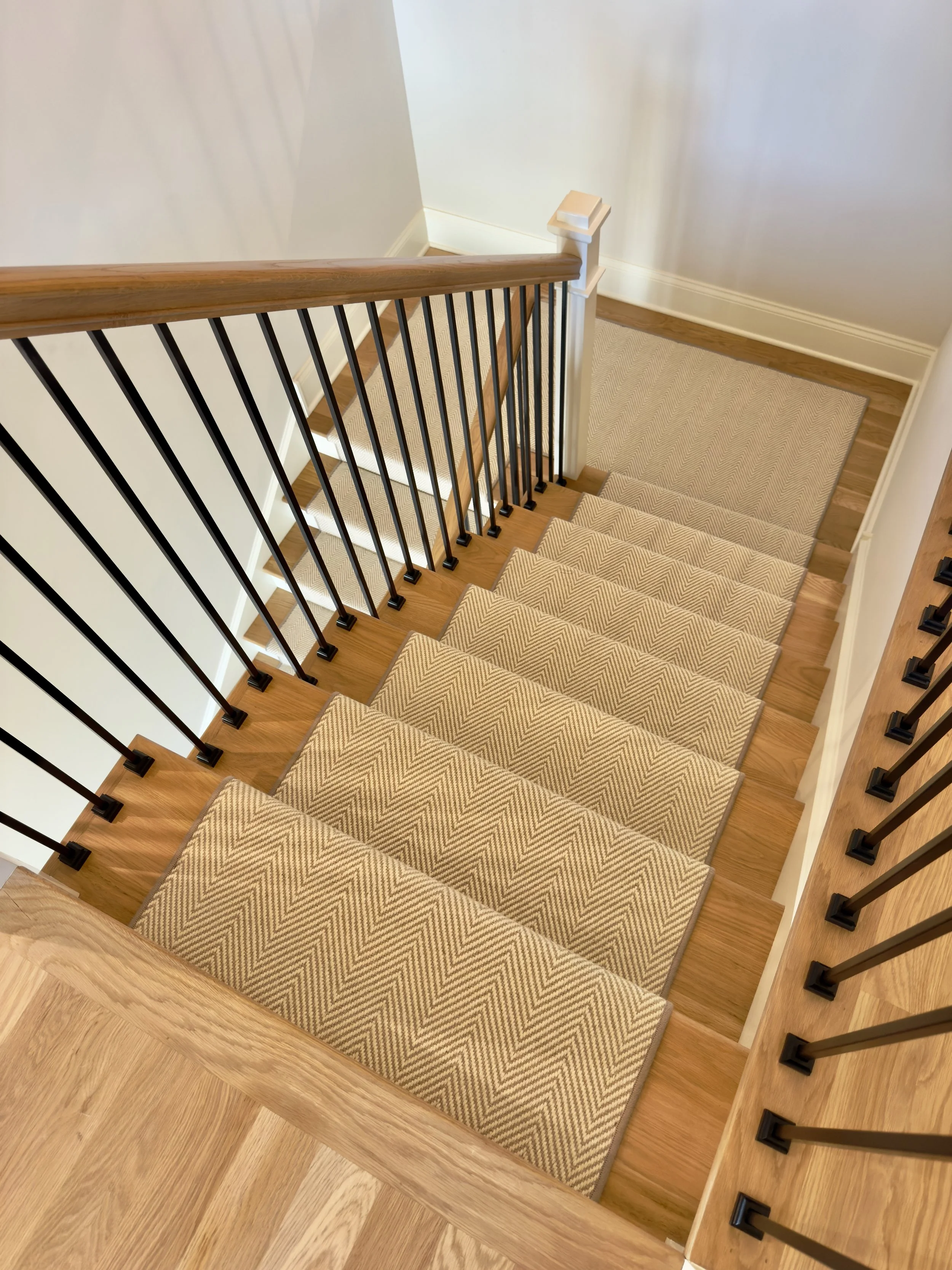 View of a wooden staircase with beige zigzag-patterned carpeted steps, black metal balusters, and a wooden handrail in a home interior.
