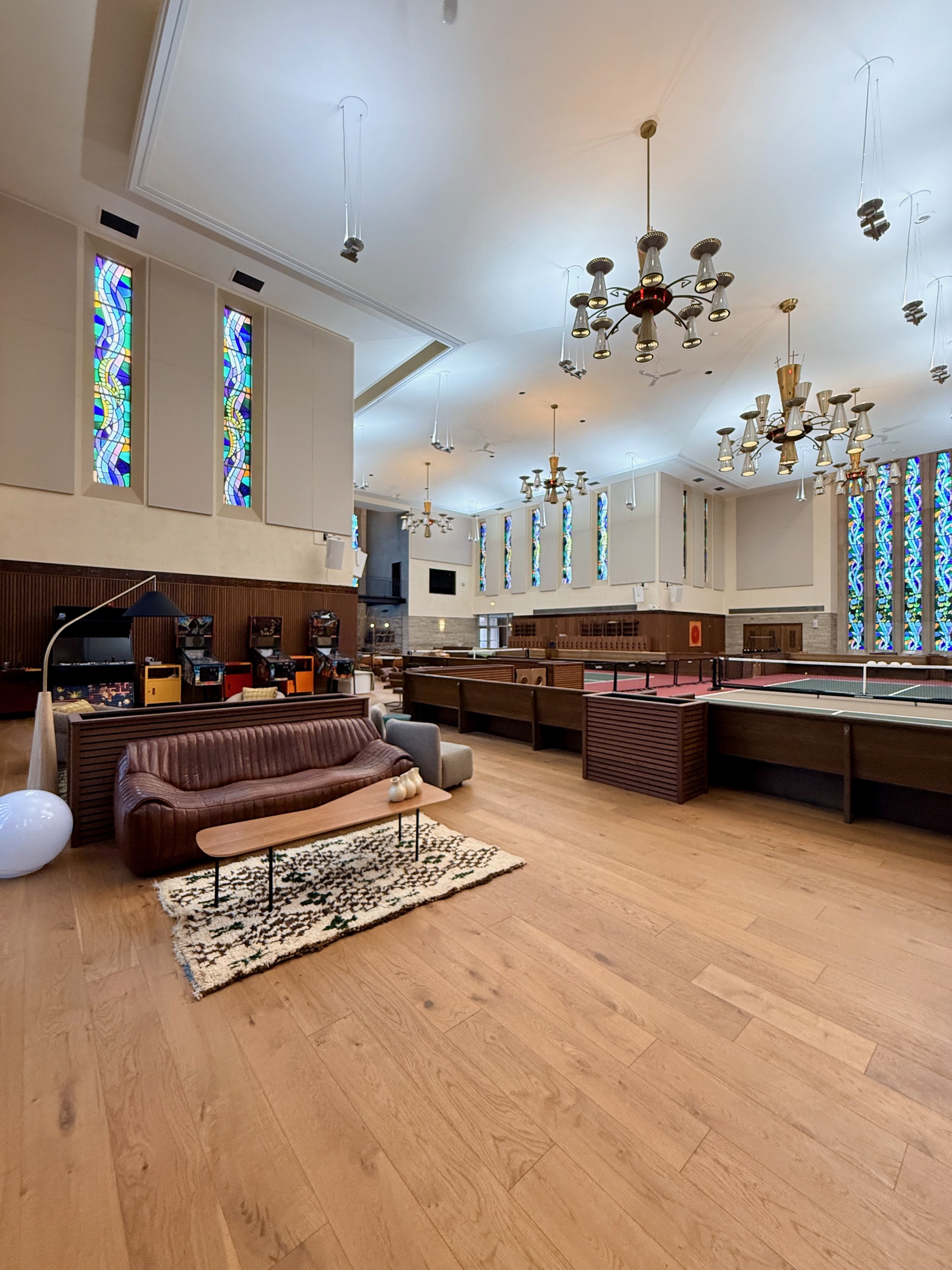 Large indoor space with wooden flooring, patterned rug, leather sofa, small coffee table, and white balloon. There are multiple chandeliers, stained glass windows, and a ping pong table.