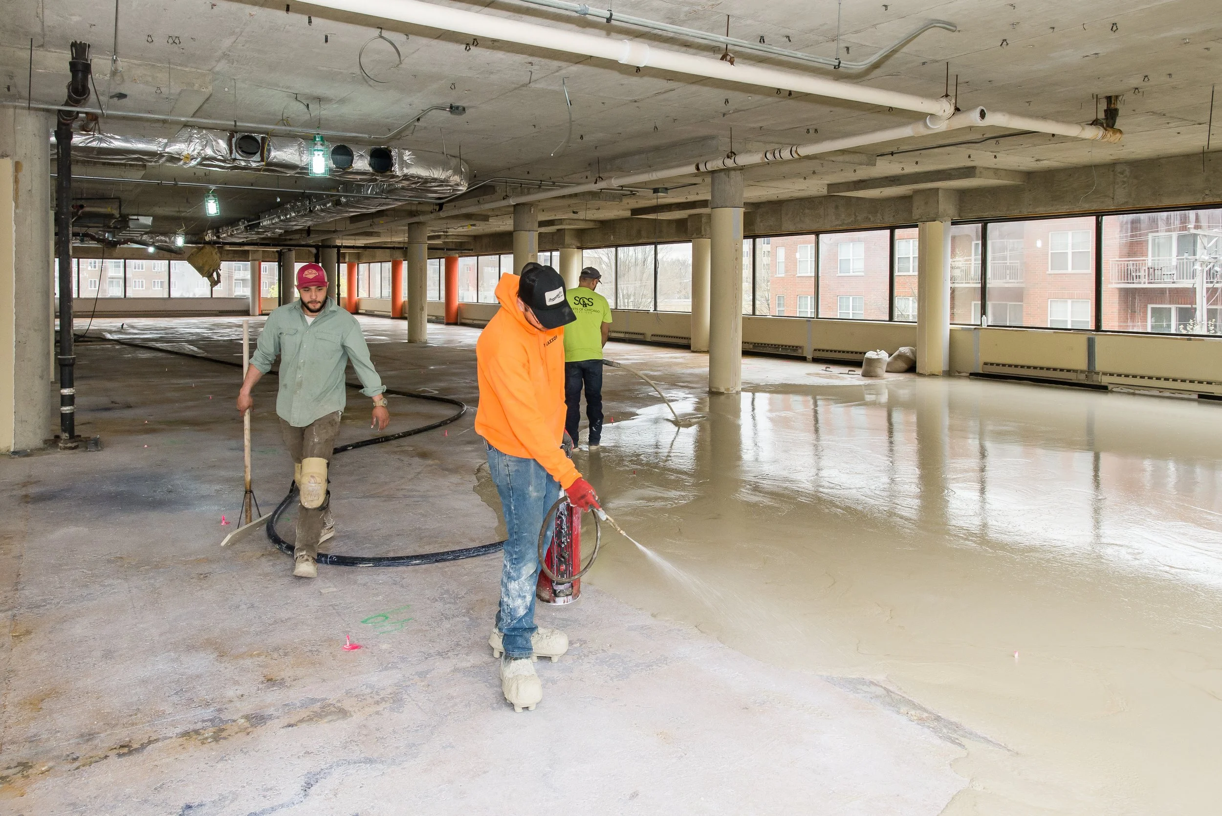 Construction workers are pouring concrete onto the floor of a building under construction, with large windows and exposed ceiling pipes.