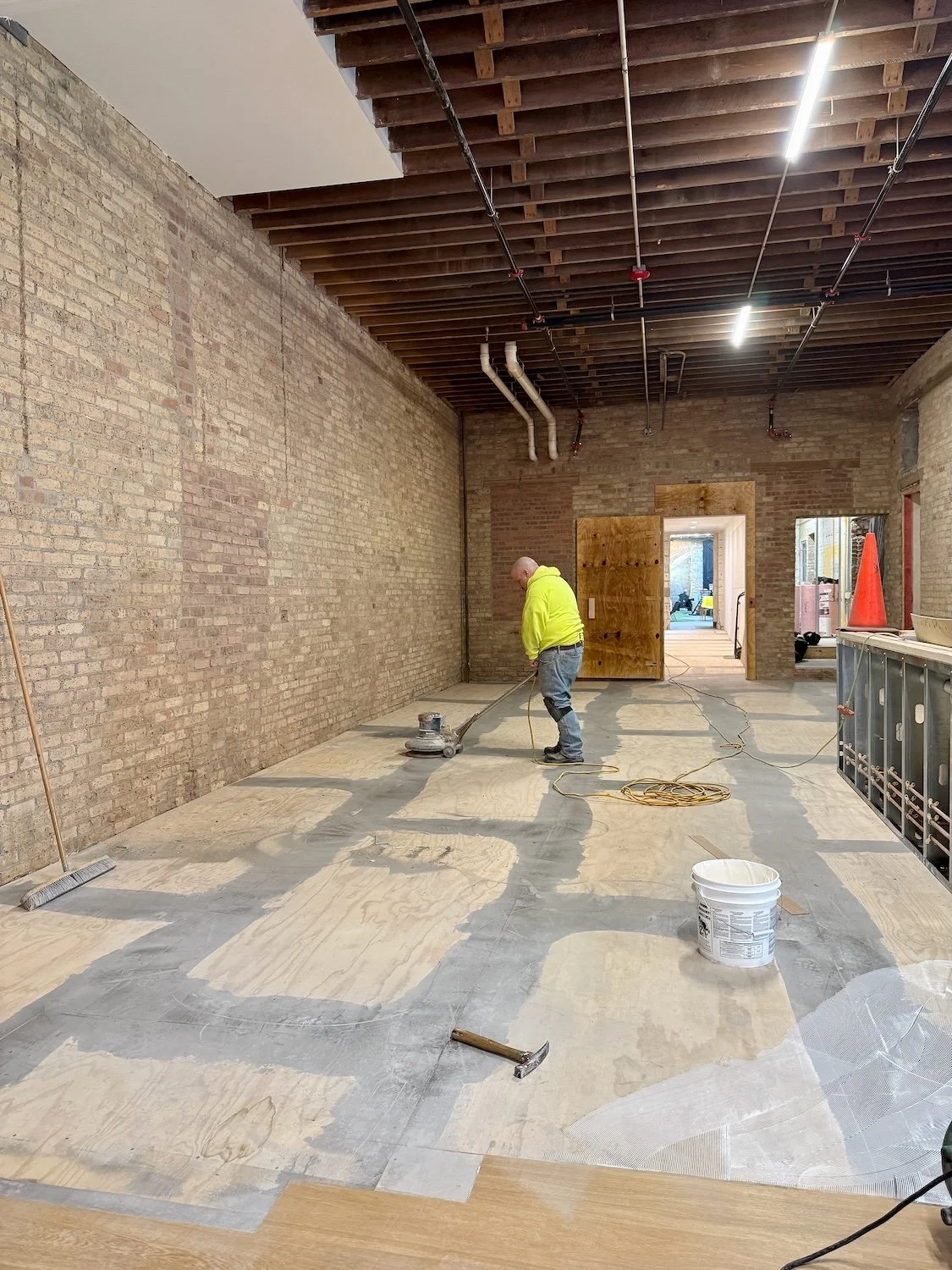 A construction worker operating a floor sander in an unfinished room with exposed brick walls and a wooden ceiling.