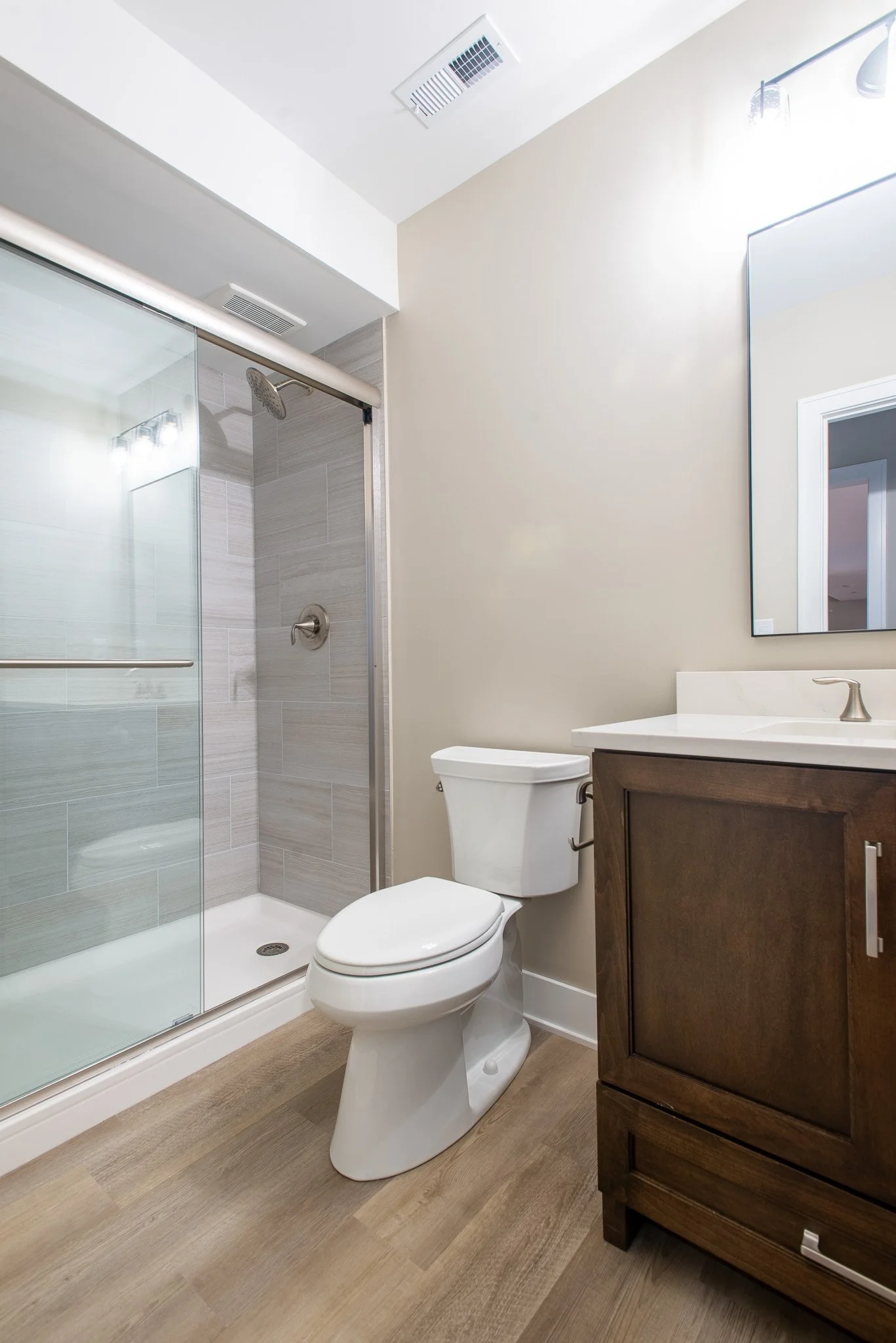 Bathroom with a glass shower enclosure, toilet, wooden vanity with a mirror, and wood-look vinyl flooring.