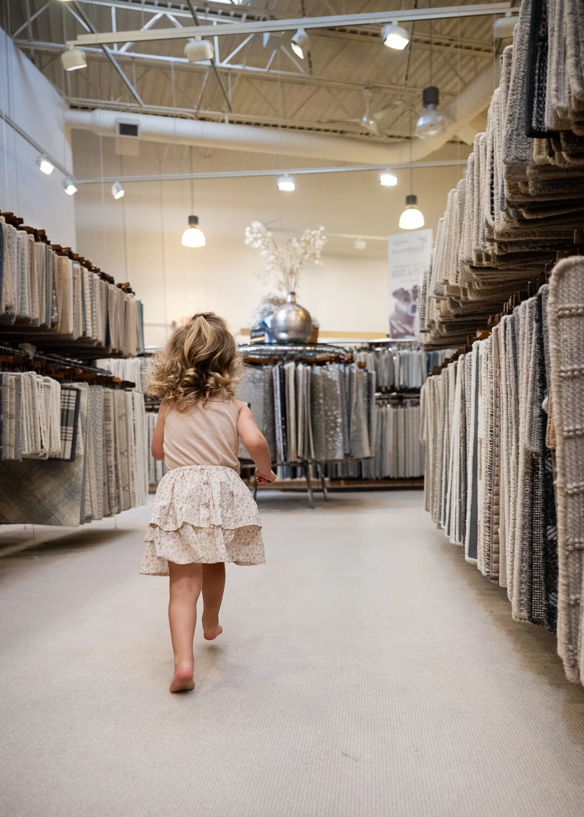 A young girl walking barefoot through a carpet store aisle lined with various carpet samples.