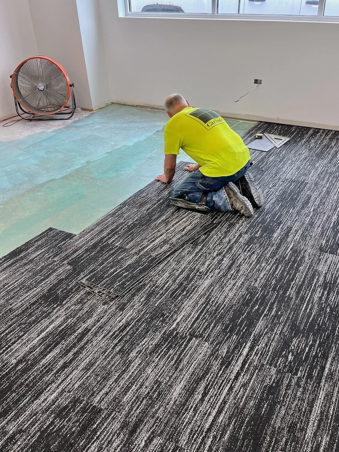 A worker kneeling on a patterned carpet in a room under installation, with a focus on the new carpet being laid and tools nearby, including a measuring tape and a square.