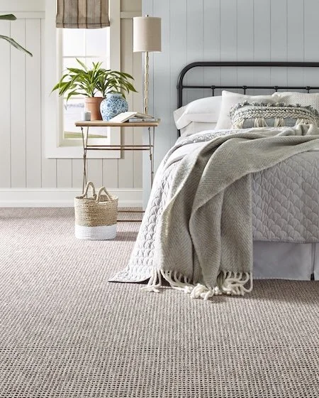 A cozy bedroom corner with a bed covered in a gray quilt and blanket, a small side table with a plant, a lamp, and an open book, with light streaming through a window.