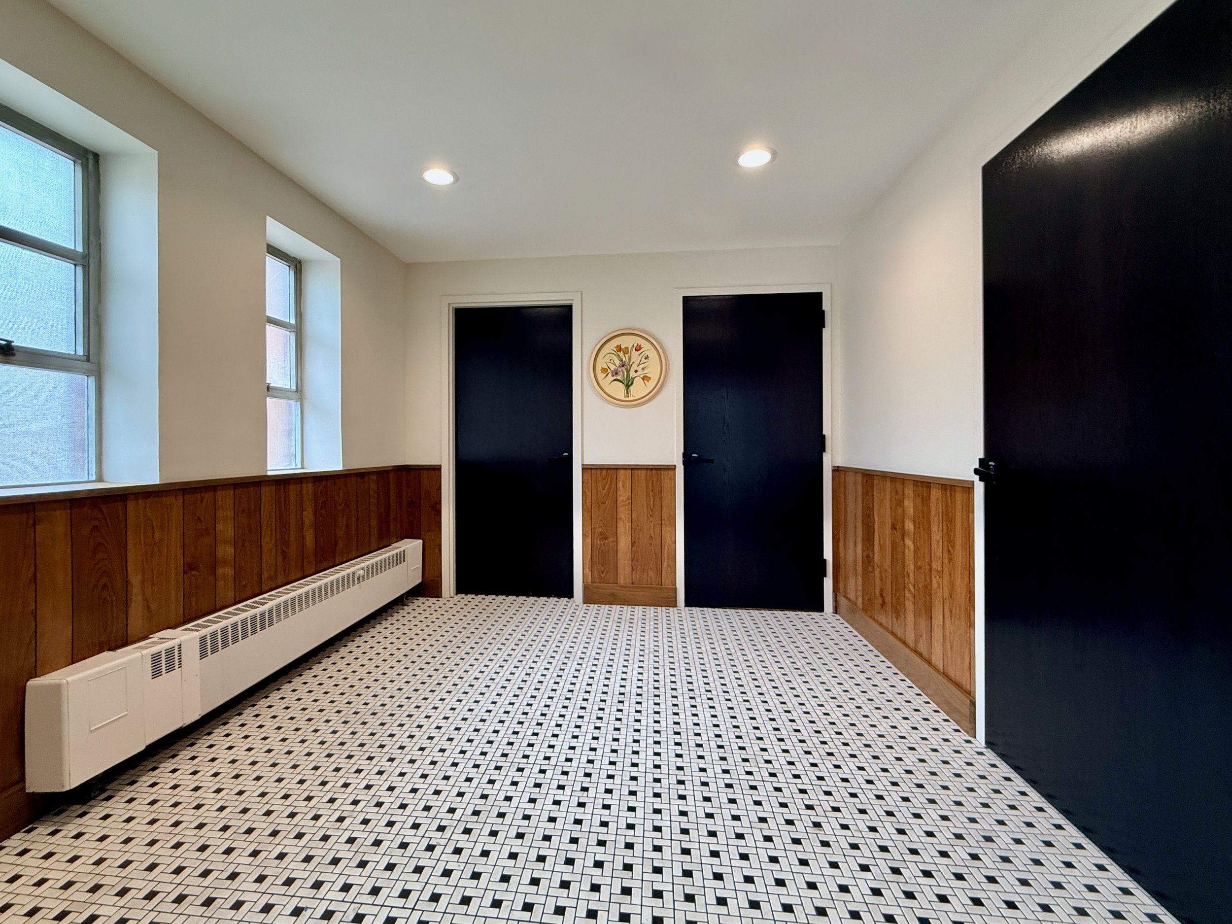 A room with white walls, three windows with blue shades, black doors, and a black and white patterned tile floor. There is wood paneling along the lower part of the walls and a circular floral artwork on the wall.