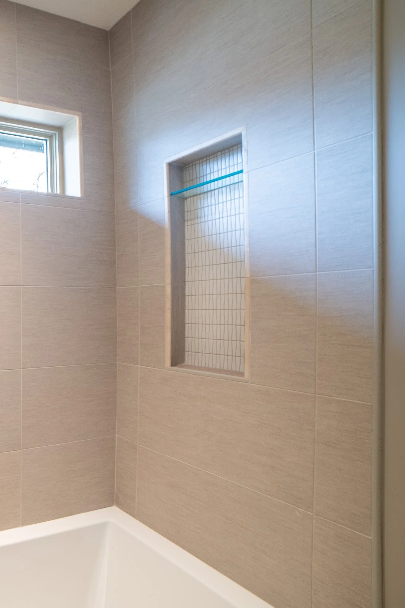 A shower with beige tiled walls, a small window near the ceiling, and a built-in niche with a glass shelf and wire grid behind it.