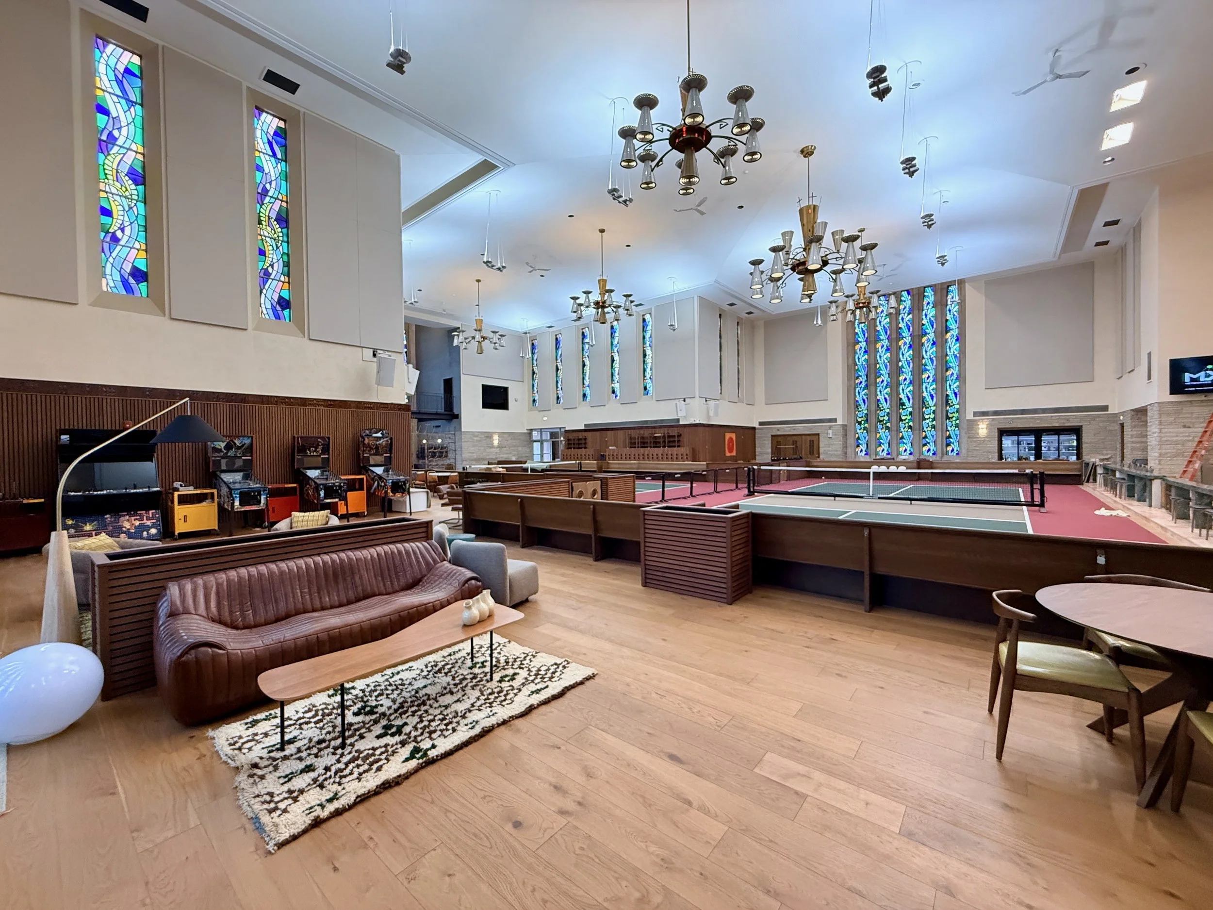 Interior of a church with stained glass windows, chandeliers, wooden pews, a ping pong table, and comfortable seating.