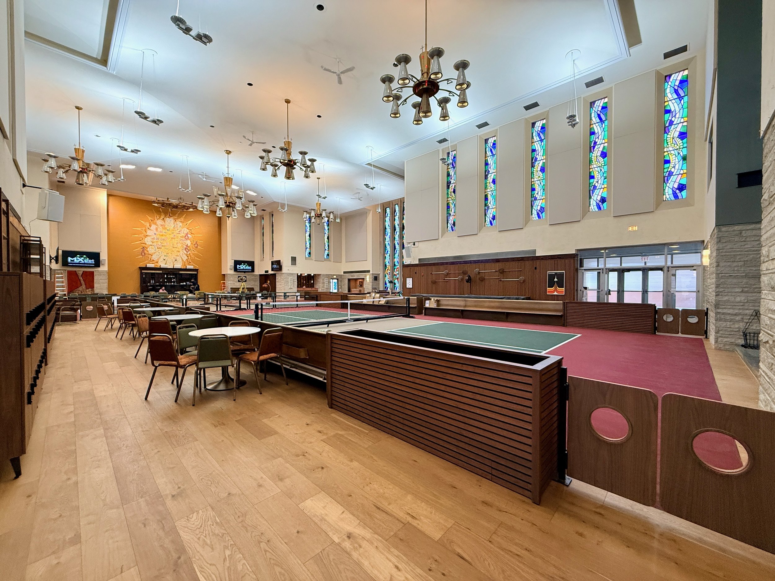Interior of a large room with a high ceiling, multiple chandeliers hanging from above, stained glass windows on the upper wall, and tables for playing ping pong.