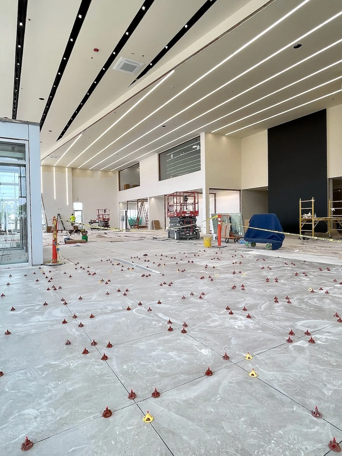 Construction site inside a building with flooring tiles being installed. Equipment such as scissor lifts, scaffolding, and tools are visible. The ceiling has modern lighting fixtures, and the space is partially finished.