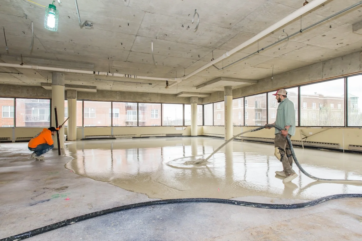 Two construction workers in a large, unfinished room are pouring and spreading a liquid concrete floor. One worker crouches near a pole on the left, and the other stands on the right holding a hose from which the concrete flows. The room has large wi