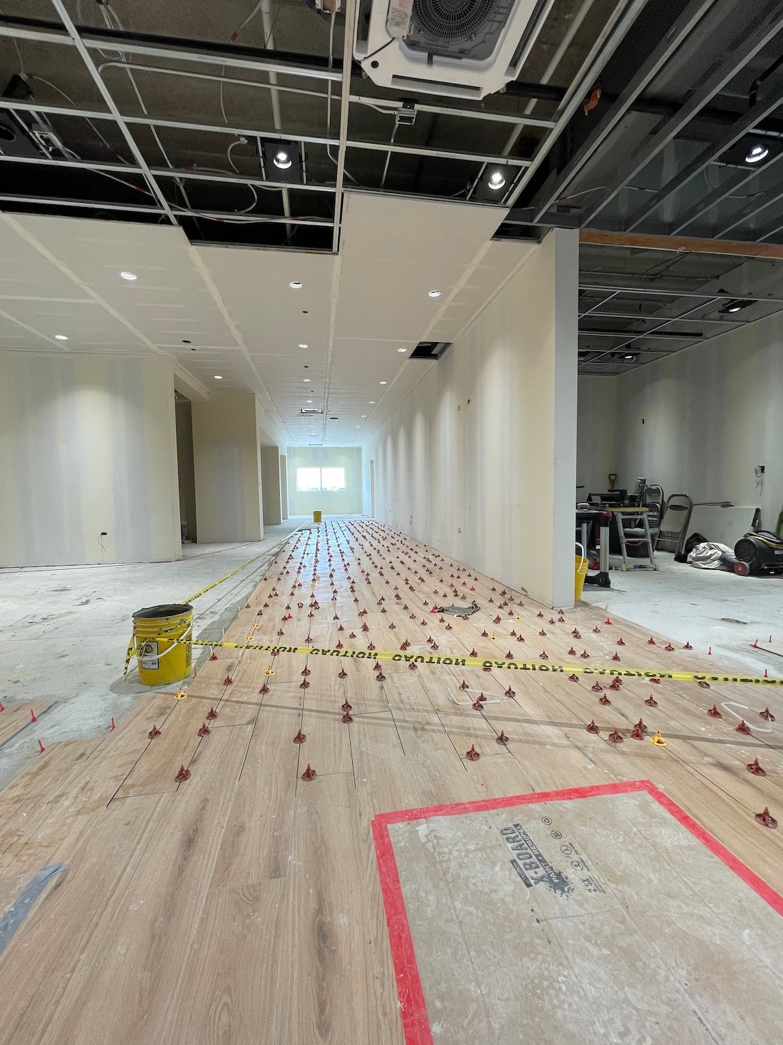 Interior of a building under construction, with a partially installed wood floor, marked with yellow caution tape and red marking, and ceiling tiles being installed.