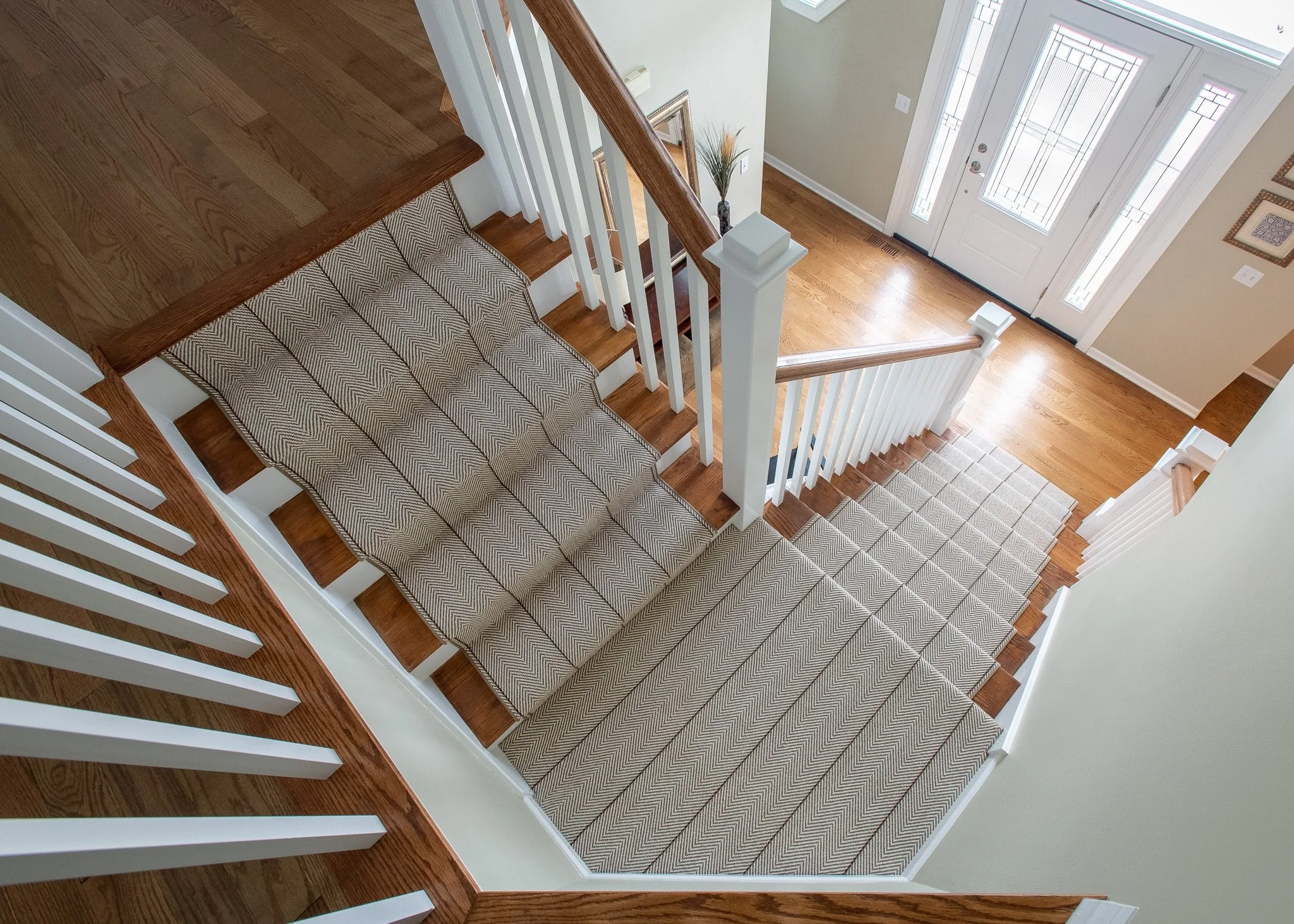 A staircase viewed from above, with wooden steps, a patterned runner, and white railings, leading to a front door with glass panels, a potted plant, and framed wall art in the entryway.