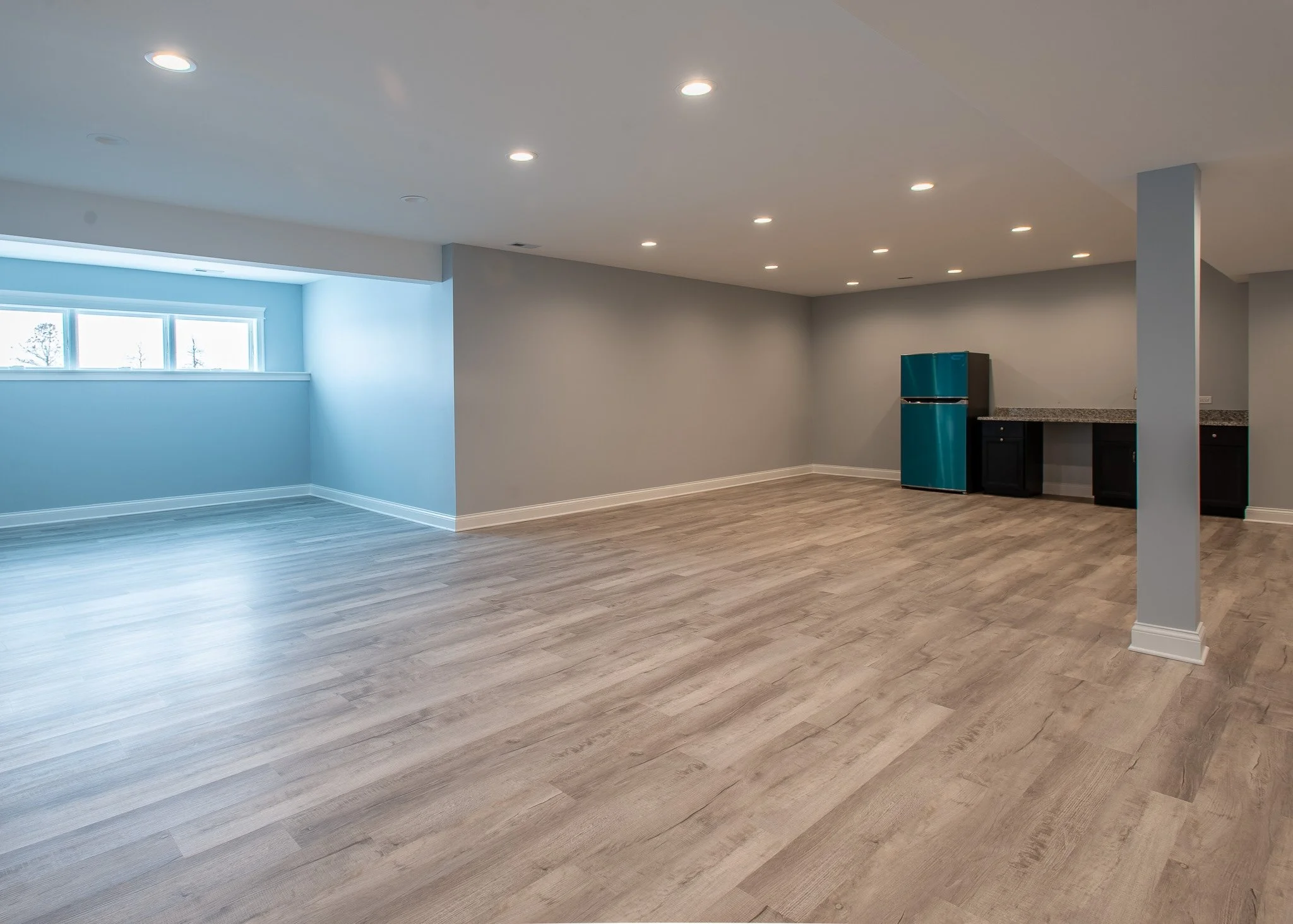 Empty basement room with beige walls, light wood flooring, a window near the ceiling, and a small refrigerator and kitchen counter in the corner.
