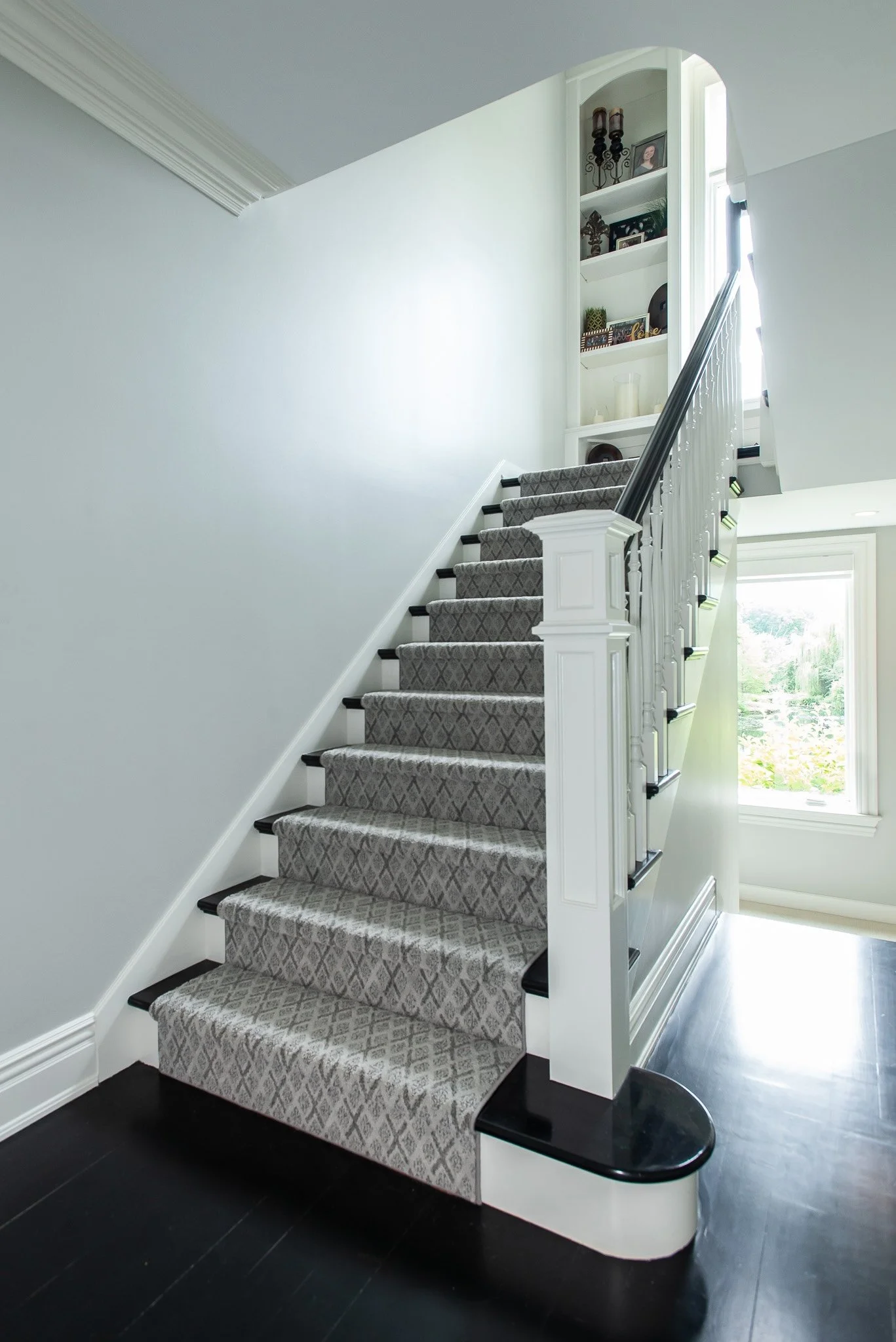 Interior view of a staircase with a grey patterned carpet, white wooden railing, and black trim. There is a window at the top of the stairs allowing natural light to illuminate the space, and built-in white shelves with decorative items on the wall b