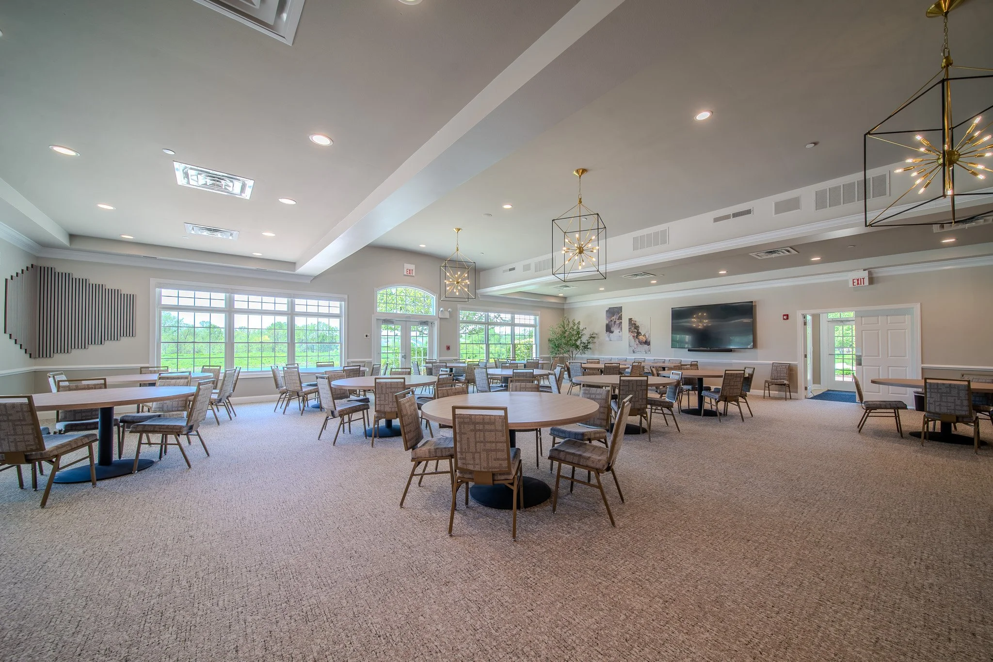 A spacious dining or event room with multiple round tables and chairs, large windows letting in natural light, modern hanging light fixtures, a flat-screen TV on the wall, and neutral-colored walls and carpeted floor.