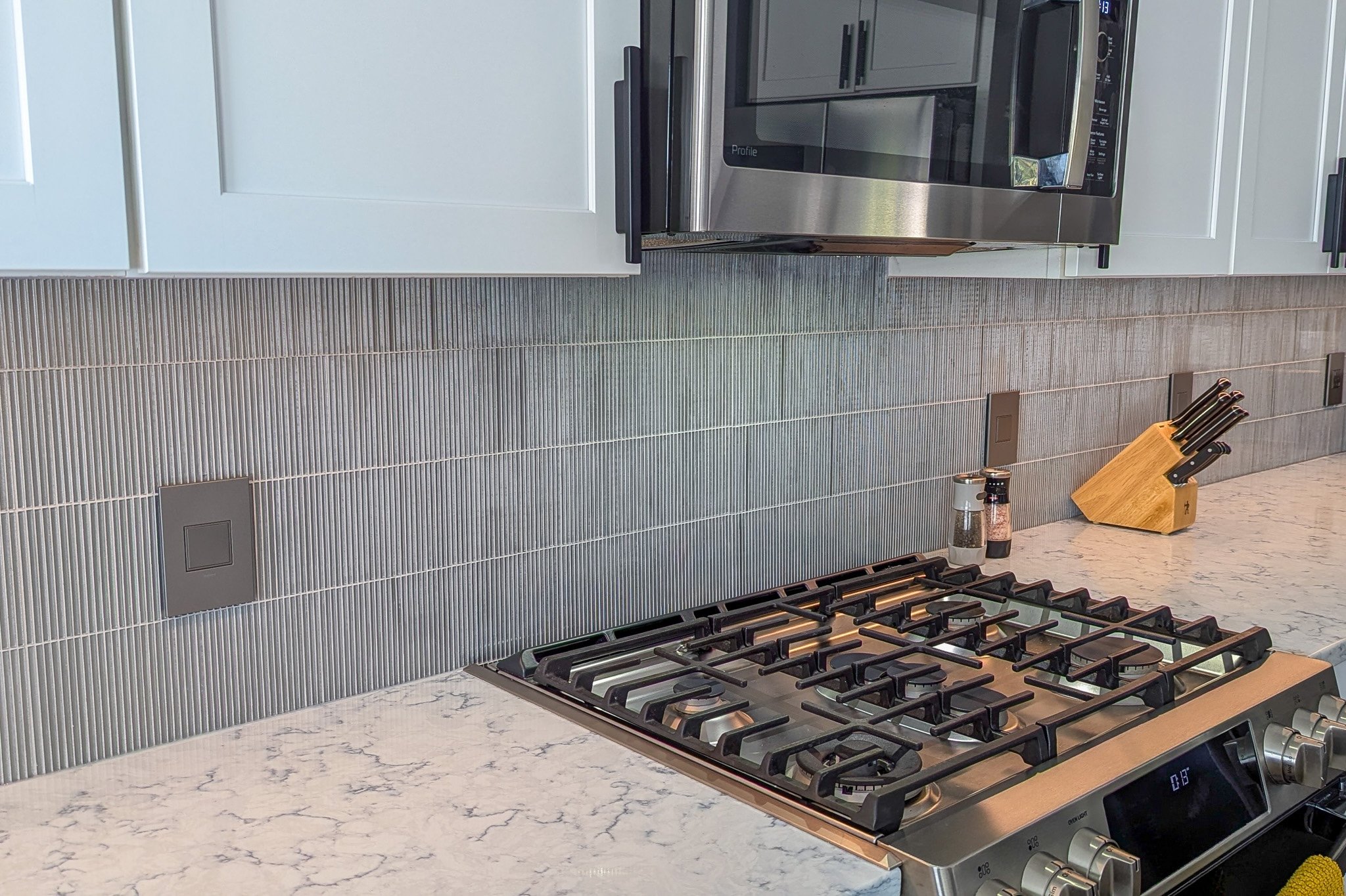 Kitchen with a stainless steel microwave oven above the stove, black stove burners, a knife block, and salt and pepper shakers on a white marble countertop. Gray textured backsplash and white cabinets.