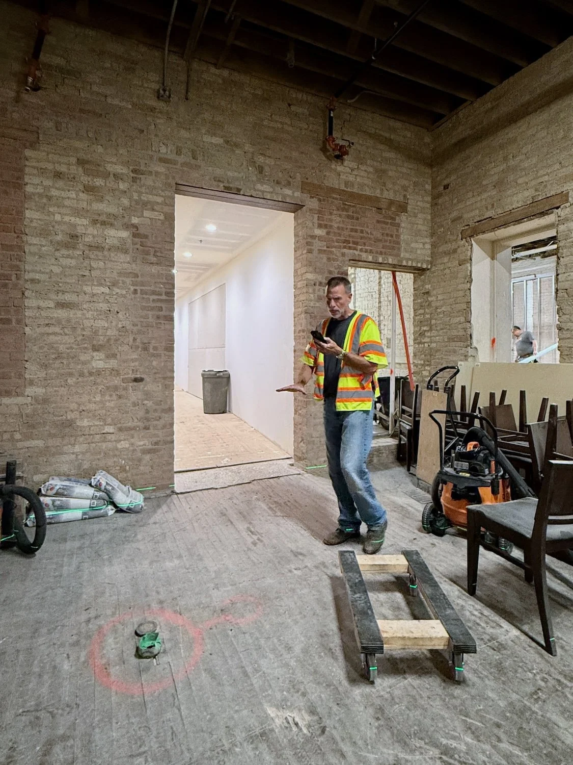 Construction worker in a high-visibility jacket using a smartphone inside a building under renovation, with exposed brick walls, construction tools, and furniture.
