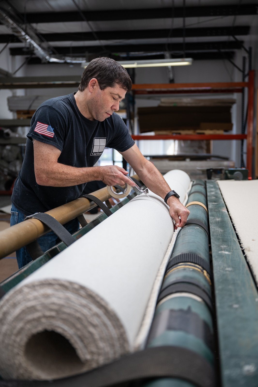 A man working in a manufacturing or workshop setting, cutting or inspecting a large roll of material, with industrial shelving and equipment in the background.