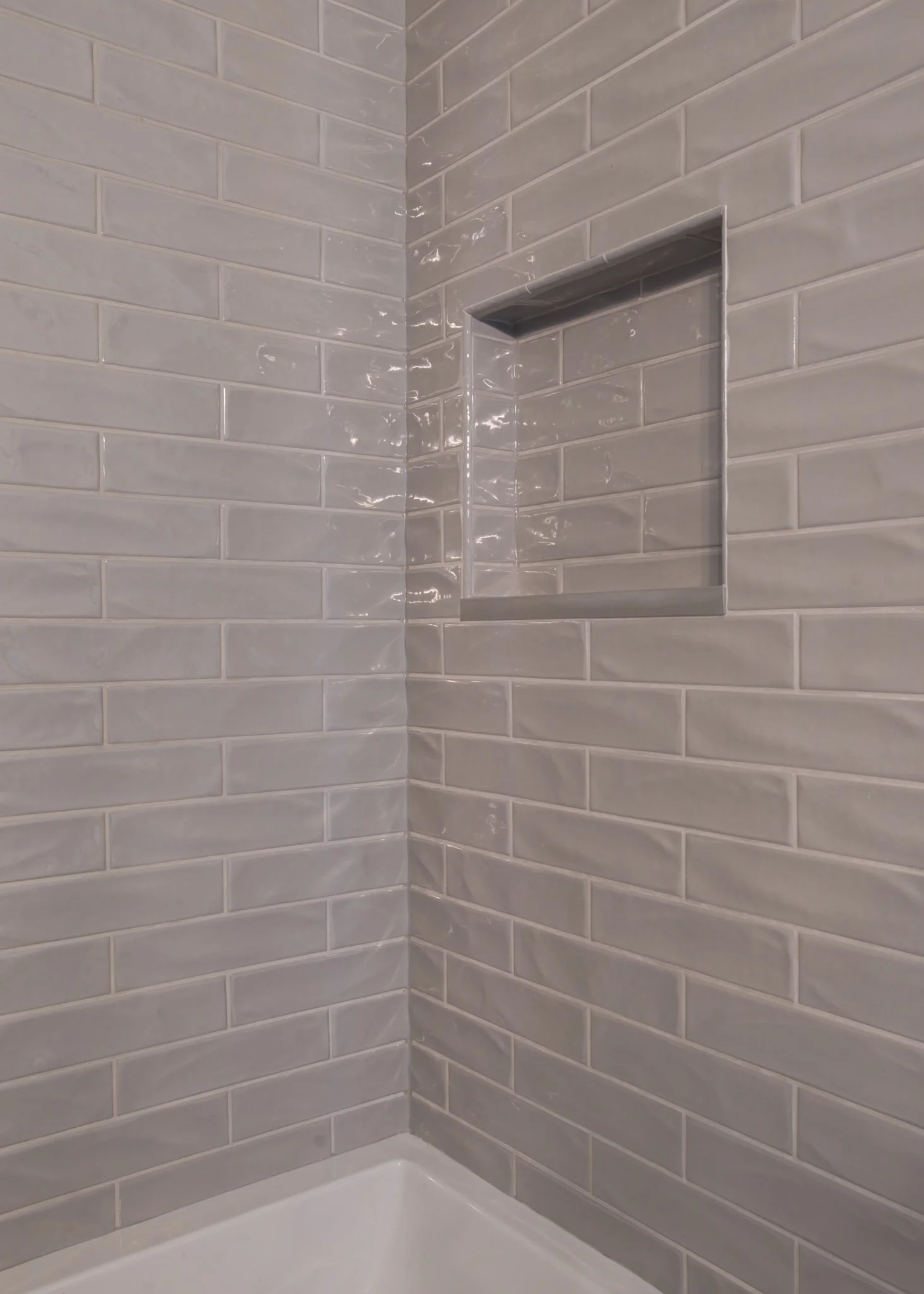 A corner of a shower with light gray subway tile walls and a built-in niche. The shower base is white, and a metal pipe runs down the wall.