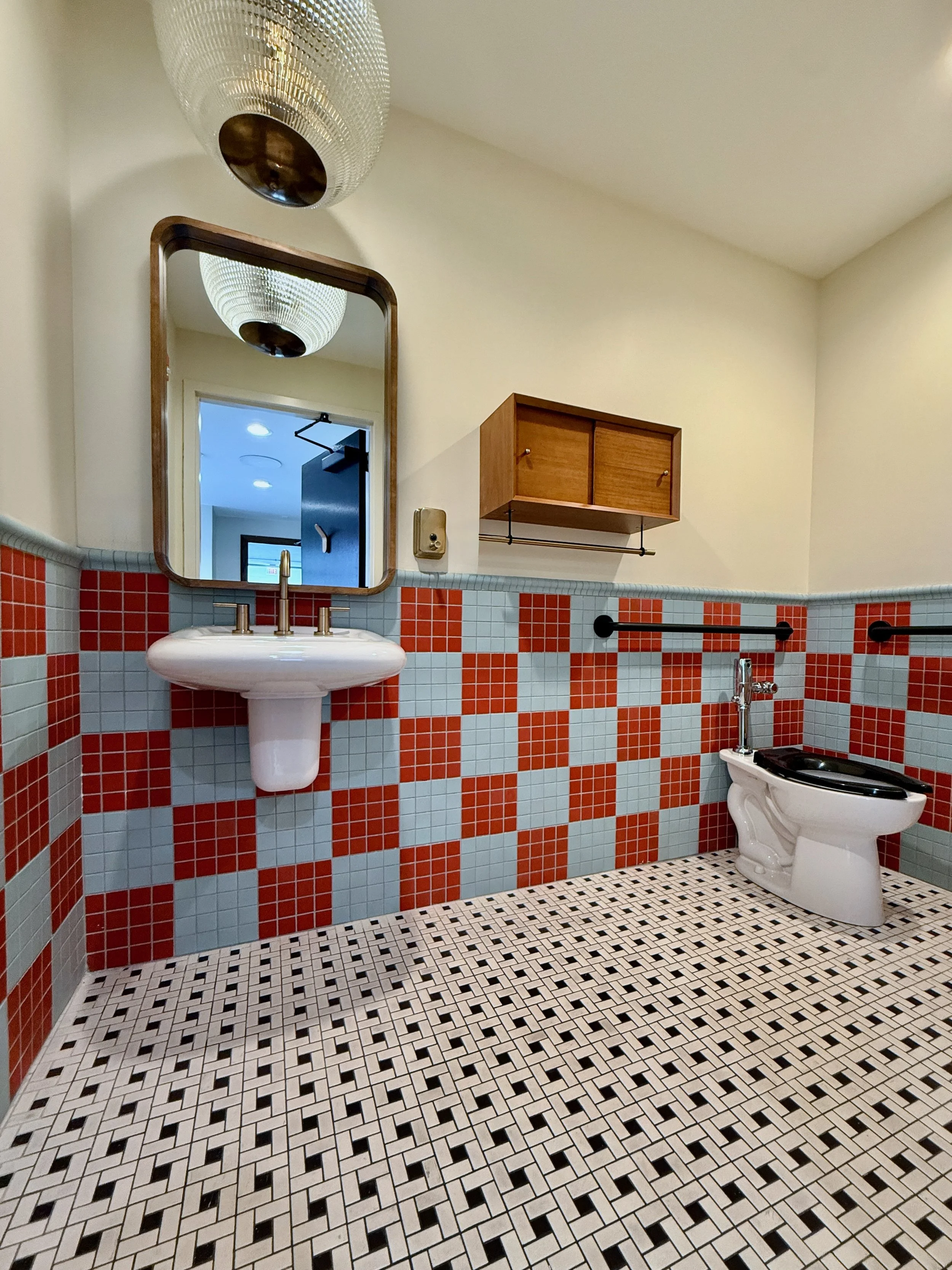 A retro-style bathroom with a black and white patterned tile floor, red and light blue checkered wall tiles, a wall-mounted mirror, a ceramic pedestal sink with a brass faucet, a wooden wall cabinet, a black towel bar, and a toilet with a black seat 