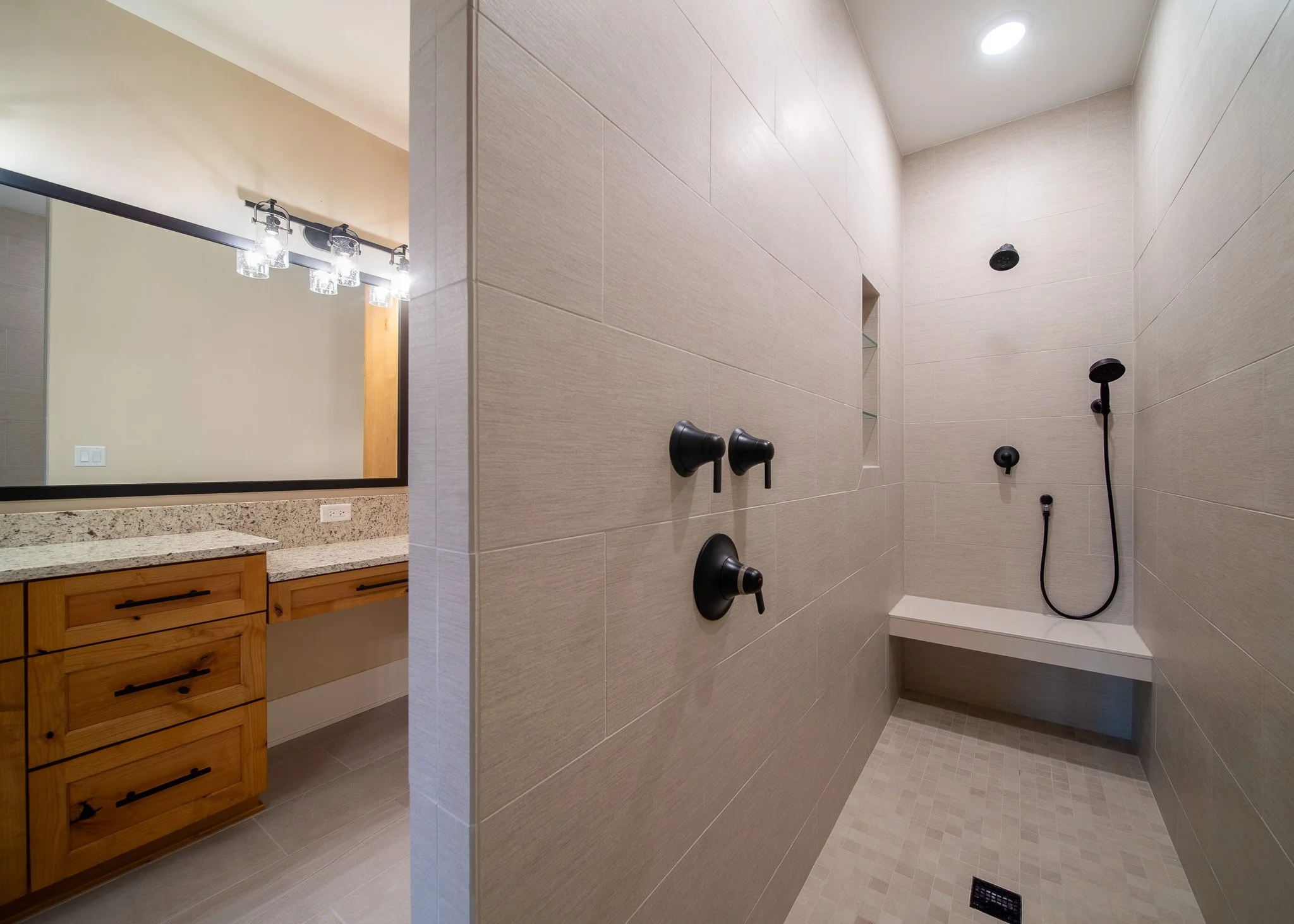 Bathroom shower area with beige tiled walls and floor, black shower fixtures, a small built-in shelf, and a ceiling light. Part of a wooden vanity with a granite countertop and a large mirror with light fixtures is visible.